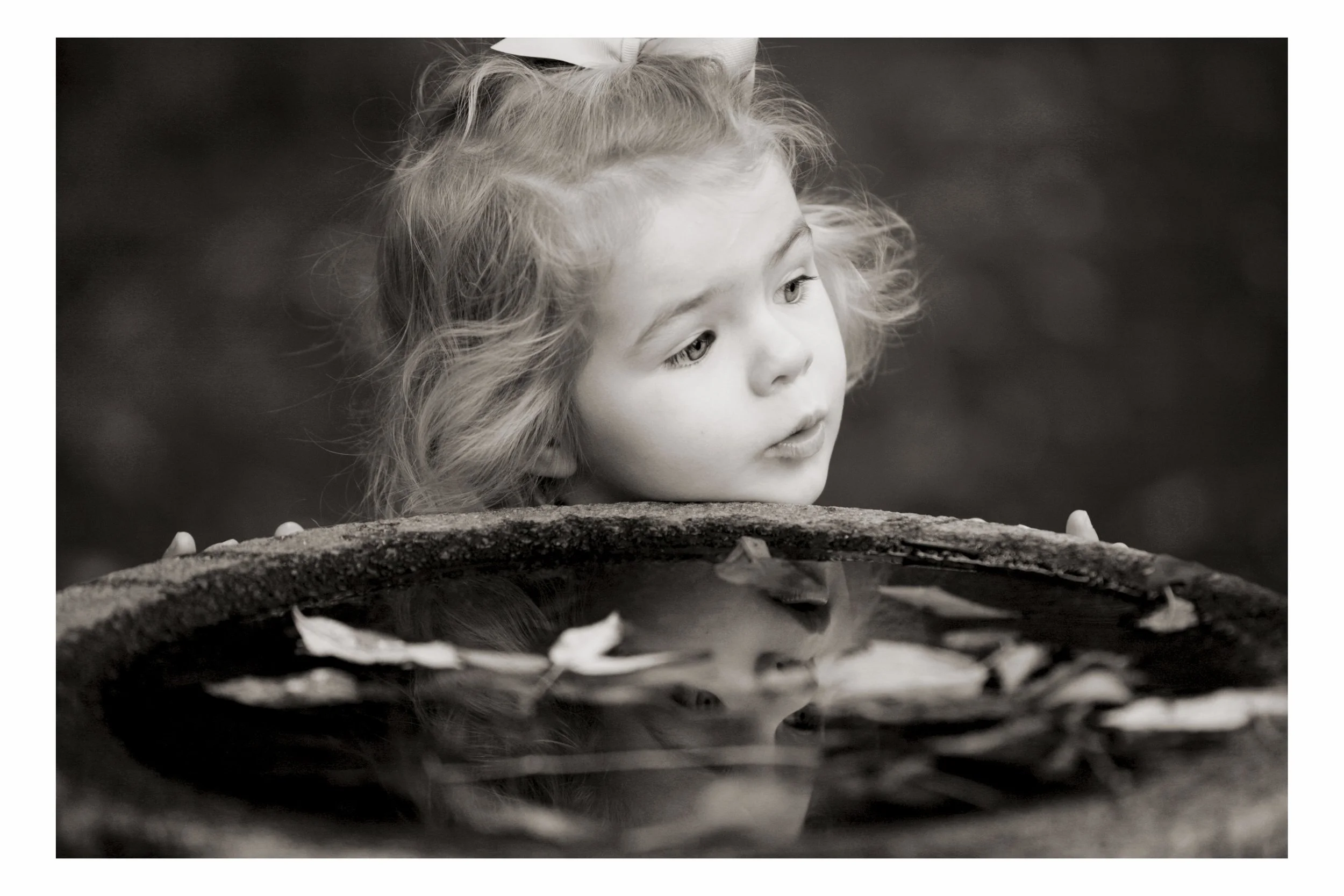 Close-up of a young girl with curly hair and a bow in her hair, looking at a pond with floating leaves.