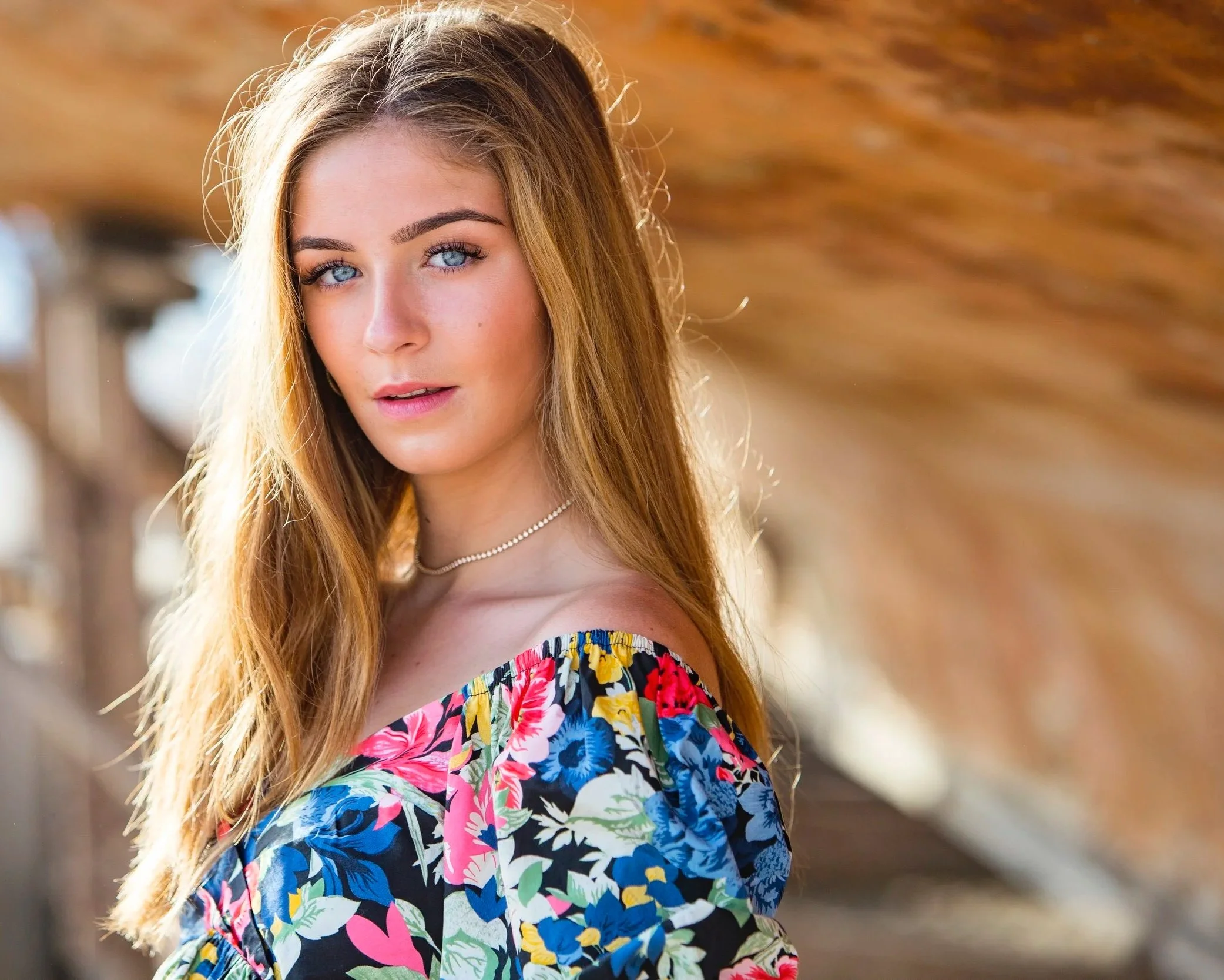 A young woman with long, wavy blonde hair and blue eyes standing outdoors, wearing a colorful off-the-shoulder floral dress. The background features a blurred rustic wooden structure.