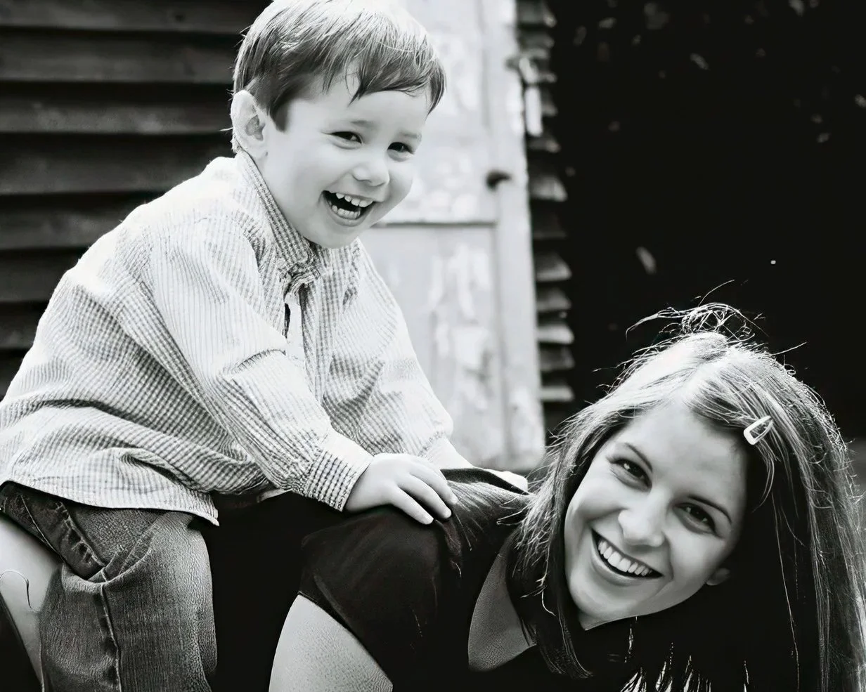 A young boy riding on a woman's back, both smiling happily outdoors.