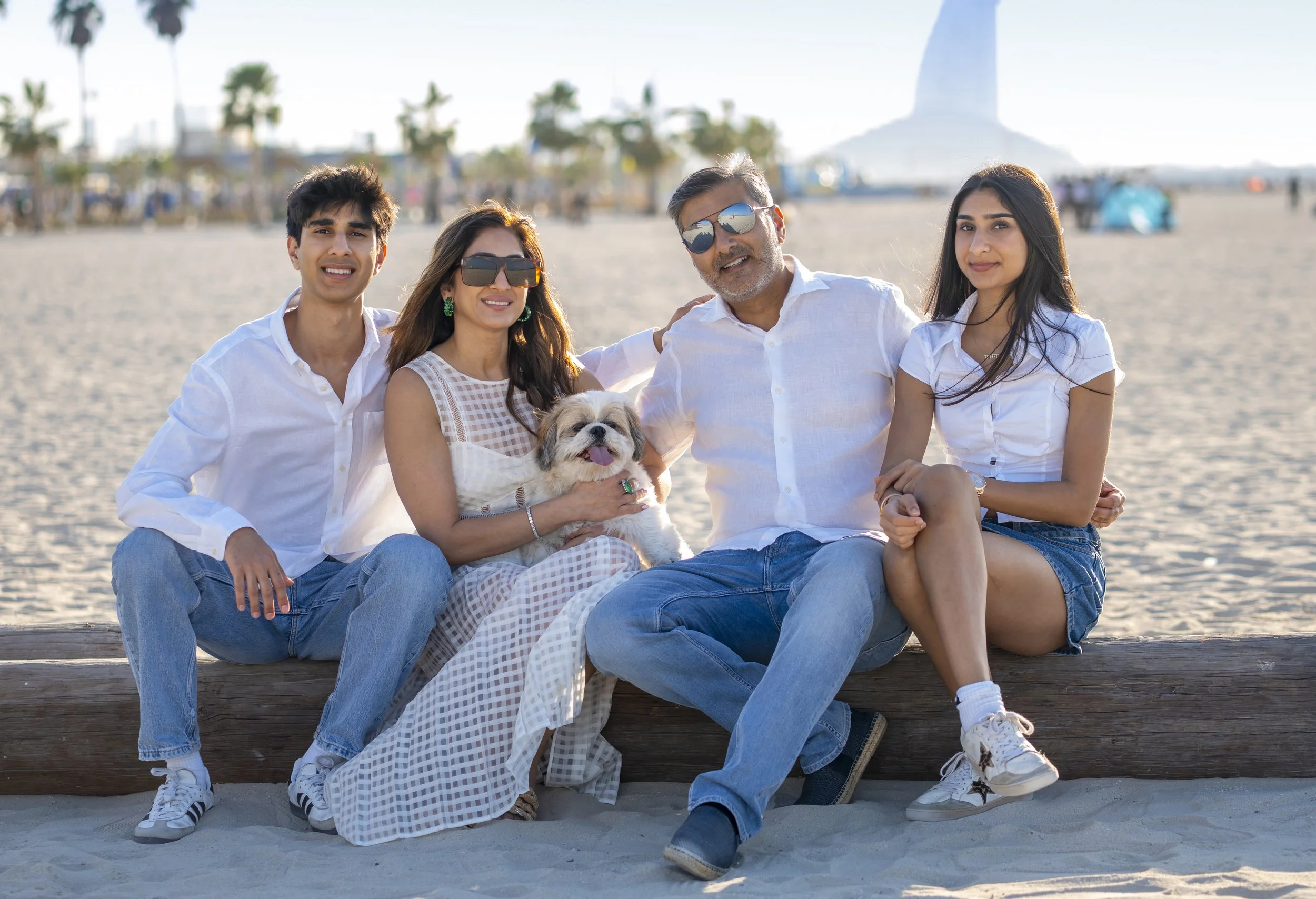 A family of five, including a dog, sitting on a wooden log at the beach with the city skyline and the statue of Christ the Redeemer in the background.