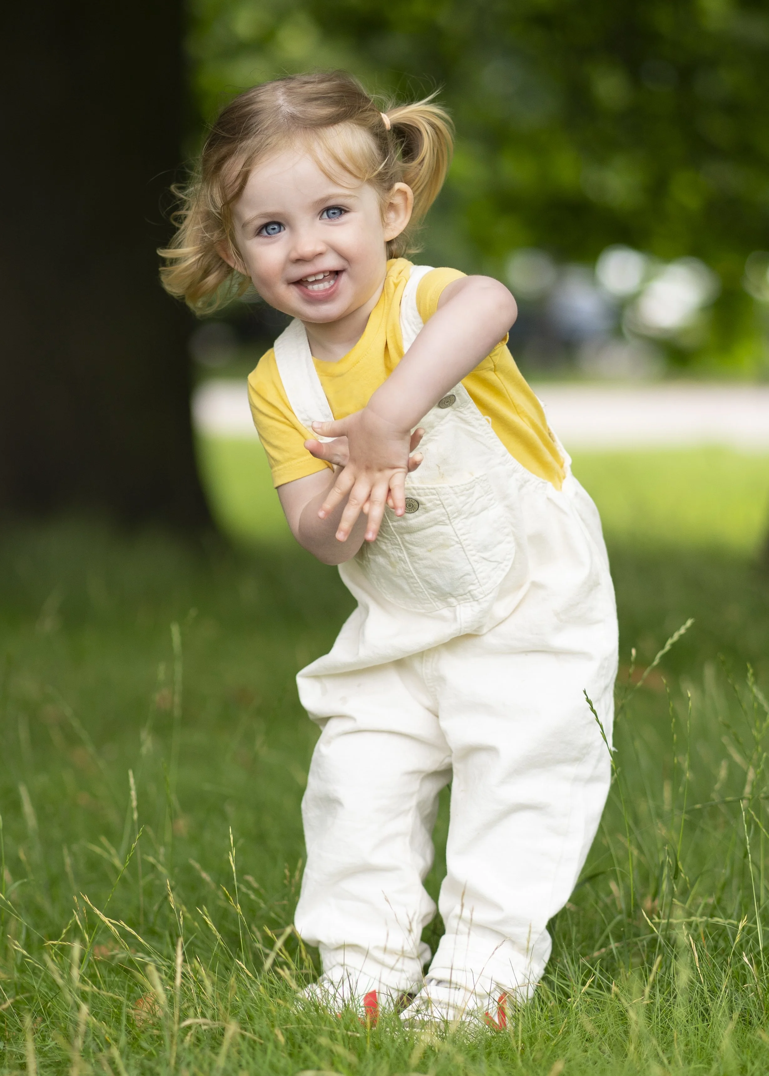 Young girl with blonde hair in pigtails, wearing a yellow t-shirt and white overalls, smiling and playing outdoors on green grass with trees in the background.