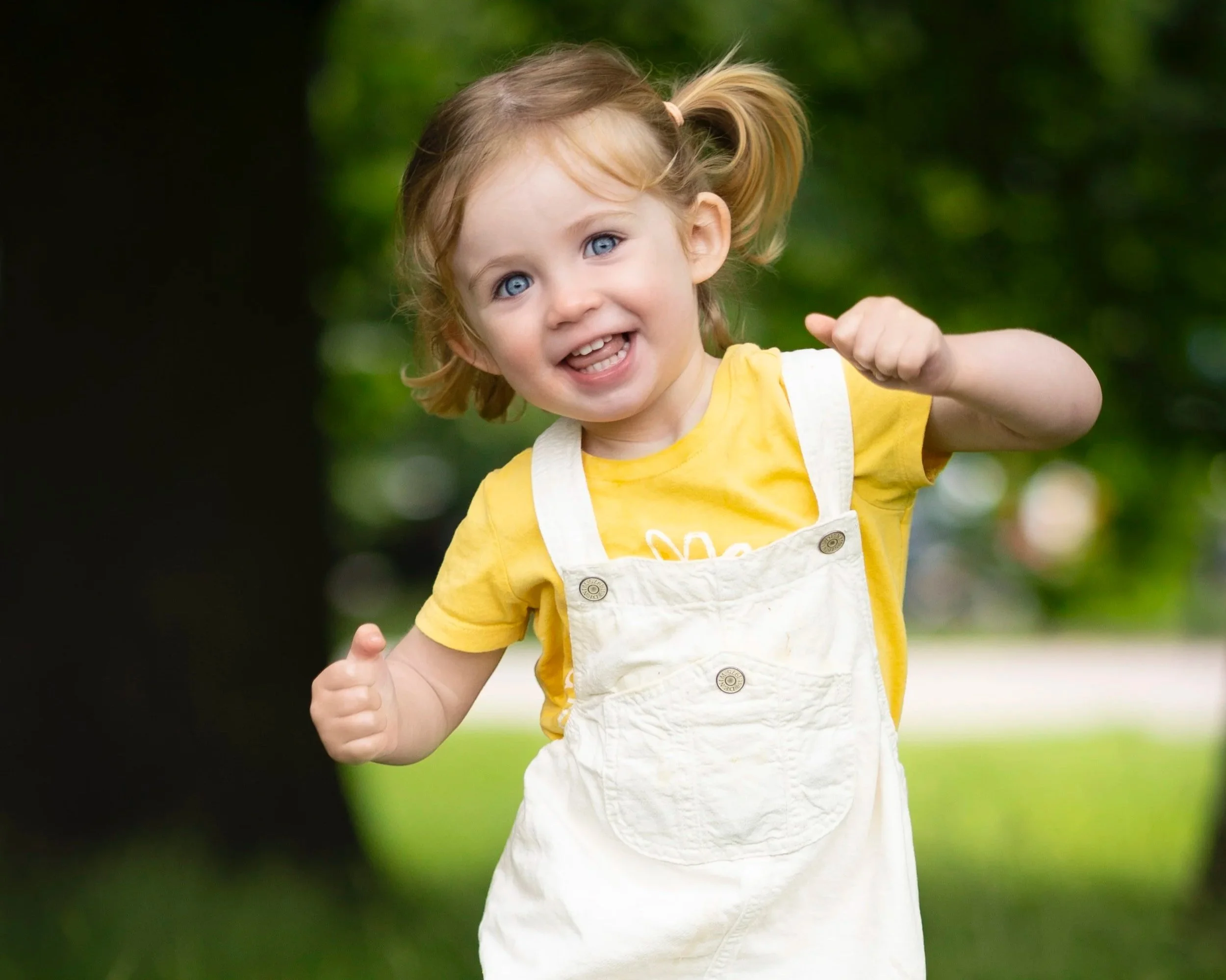 A young girl with blue eyes and blonde hair tied in pigtails, wearing a yellow shirt and white overalls, smiling and running outdoors in a park with green trees in the background.