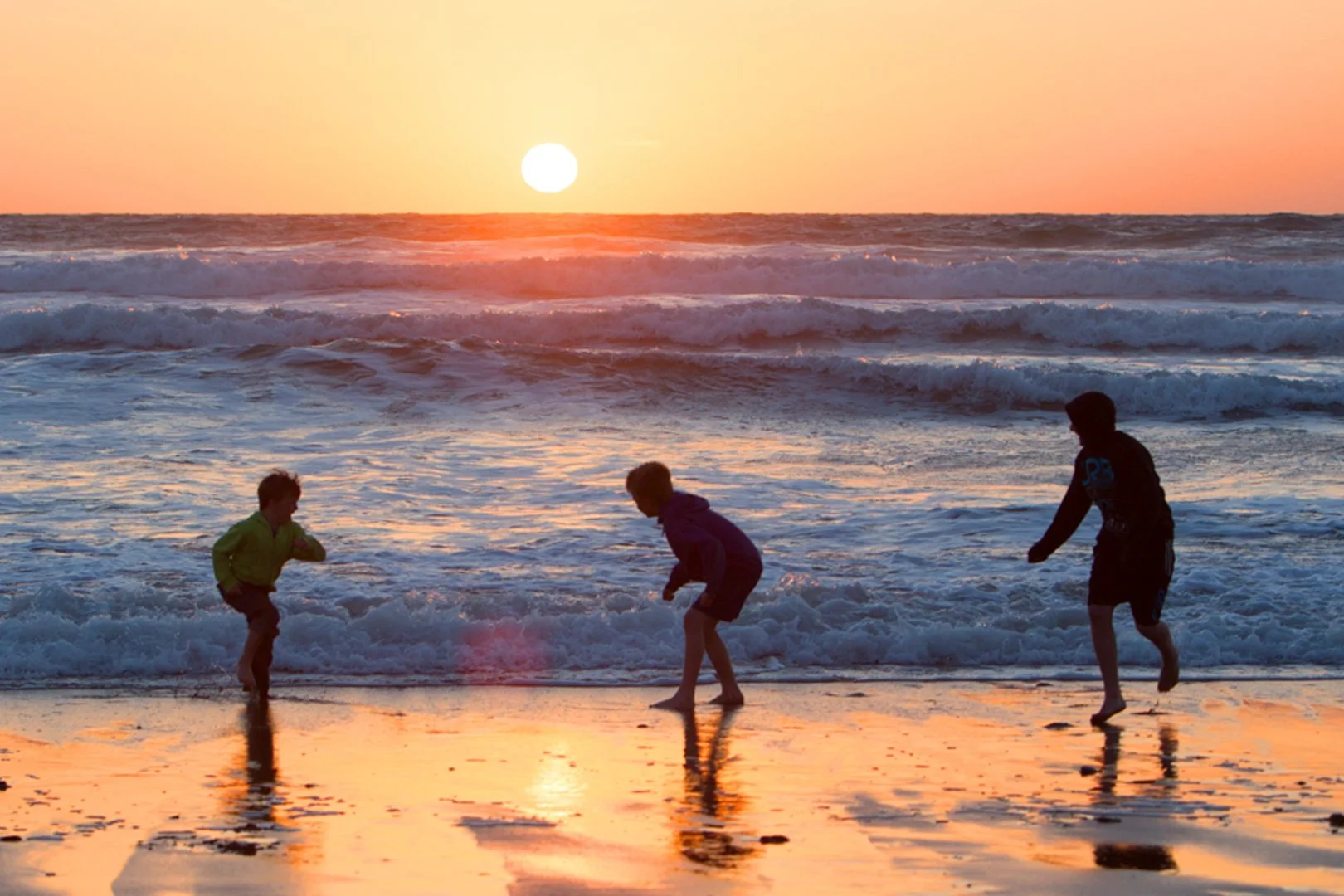 Three children playing in the surf at sunset on a beach, with the sun low on the horizon and casting a warm glow over the water and sand.