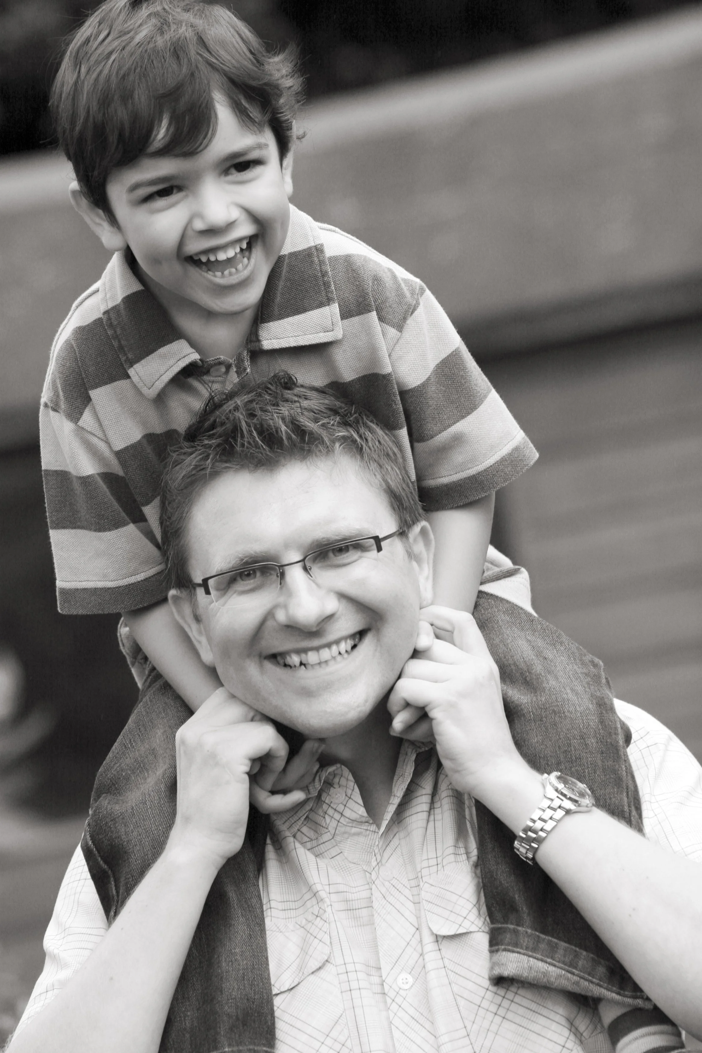A man with glasses and a watch carrying a young boy on his shoulders, both smiling and appearing joyful in an outdoor setting.