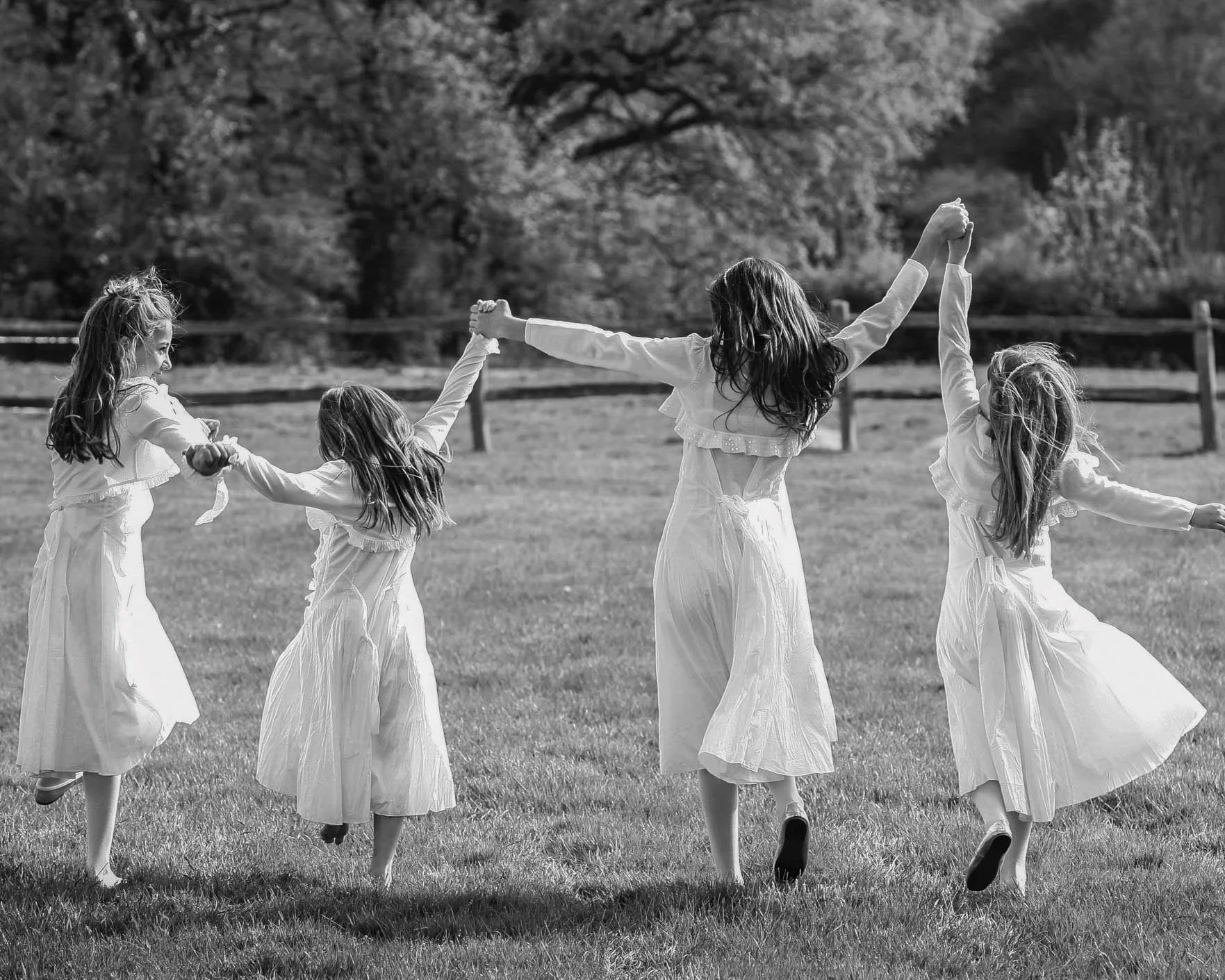 Four young girls in white dresses holding hands and dancing in a grassy field, with trees and a wooden fence in the background.