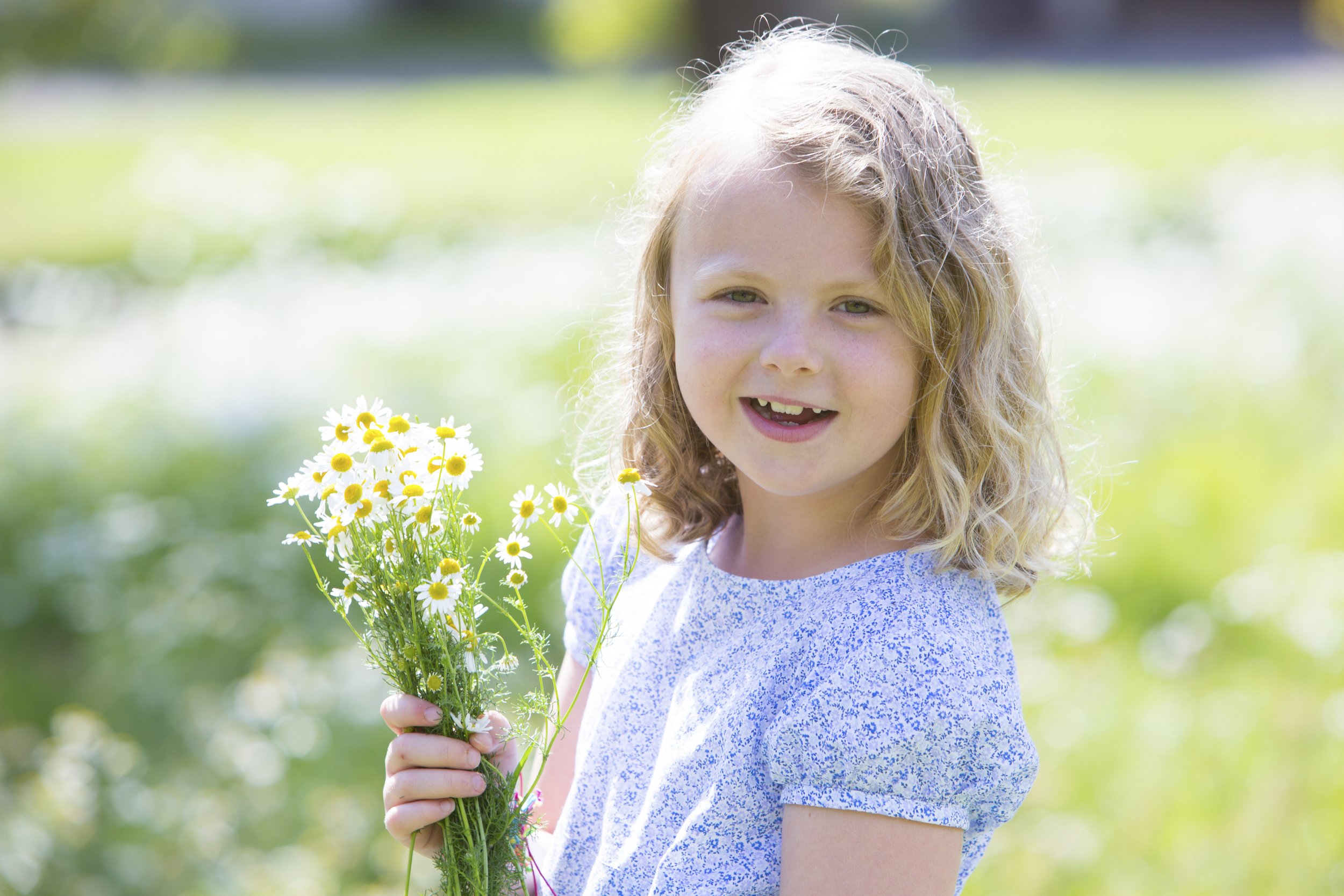 A young girl with curly blonde hair holding a bouquet of daisies outdoors on a sunny day.