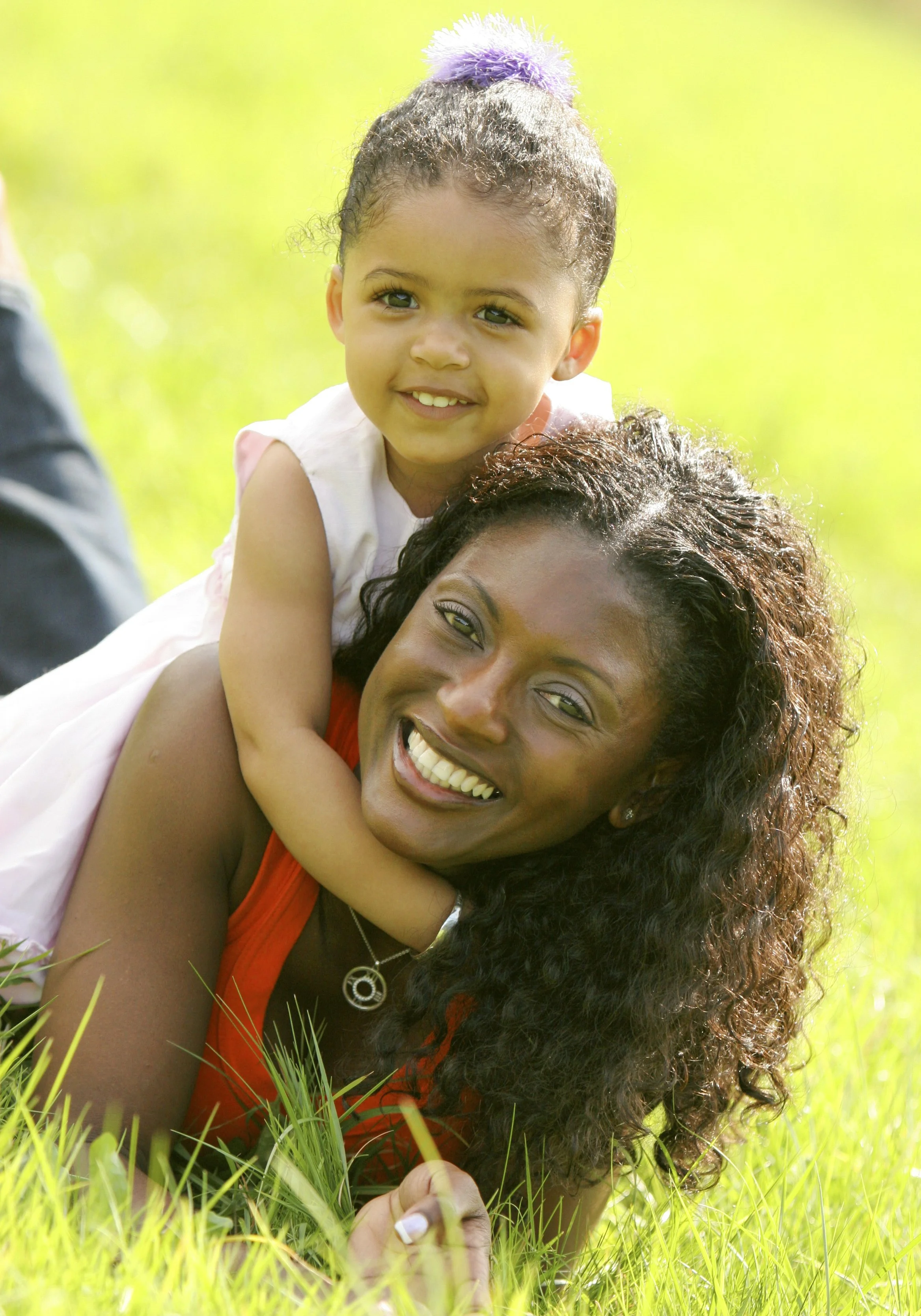 A woman with curly dark hair lying on the grass smiling while a little girl with braided hair and a purple hair tie peers over her shoulder, both outdoors on a sunny day.