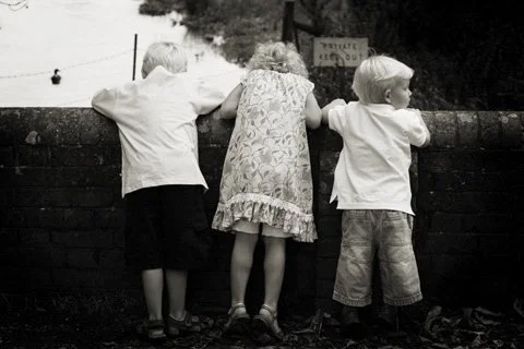 Three children standing on a wall, looking over a river or lake, with their backs facing the camera.