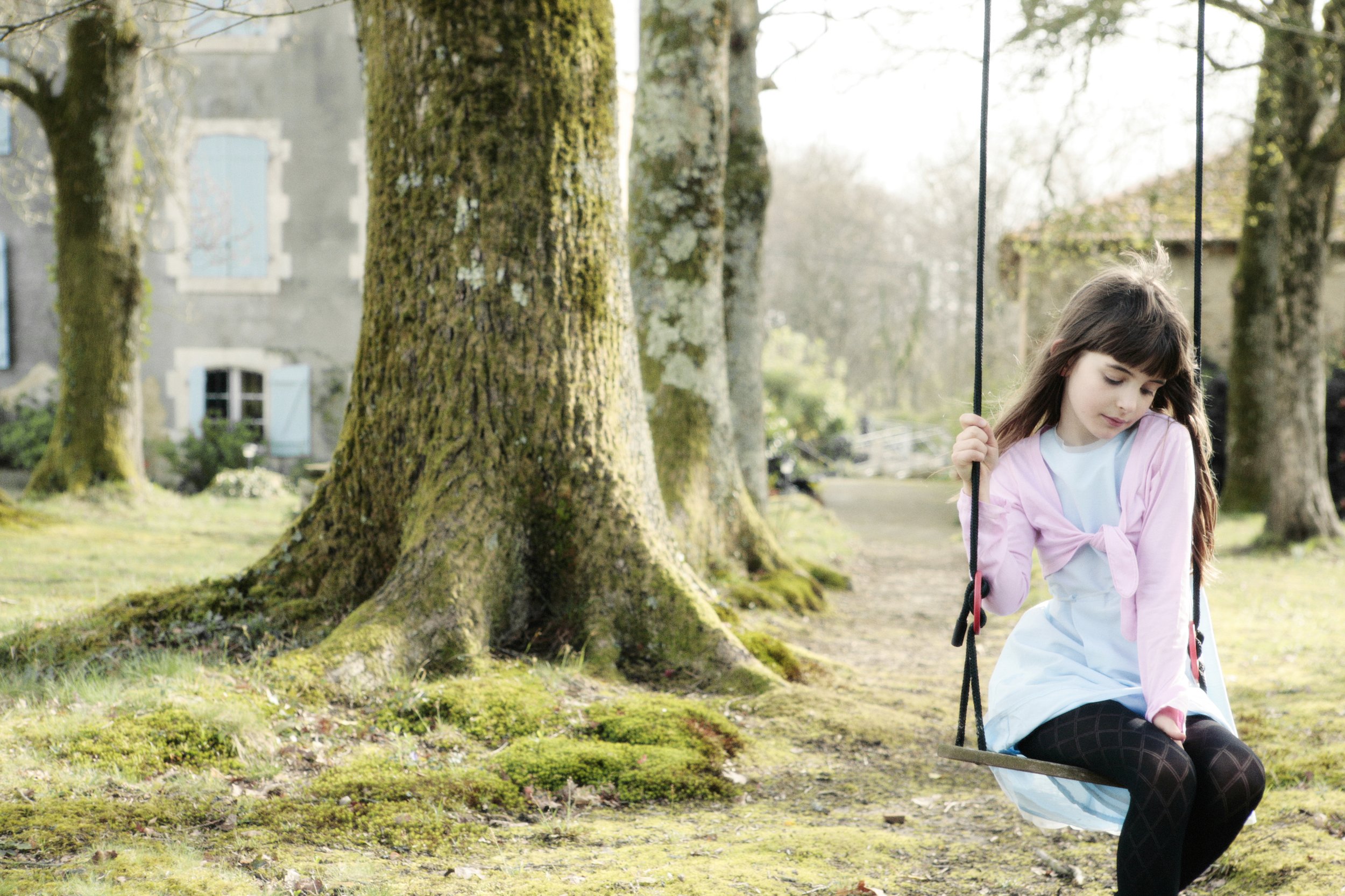 A young girl with dark hair sitting on a swing in a park with large trees and a building in the background.