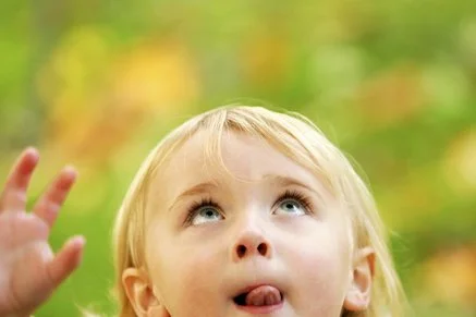 A young girl with blonde hair looking up outdoors with trees and colorful leaves in the background.
