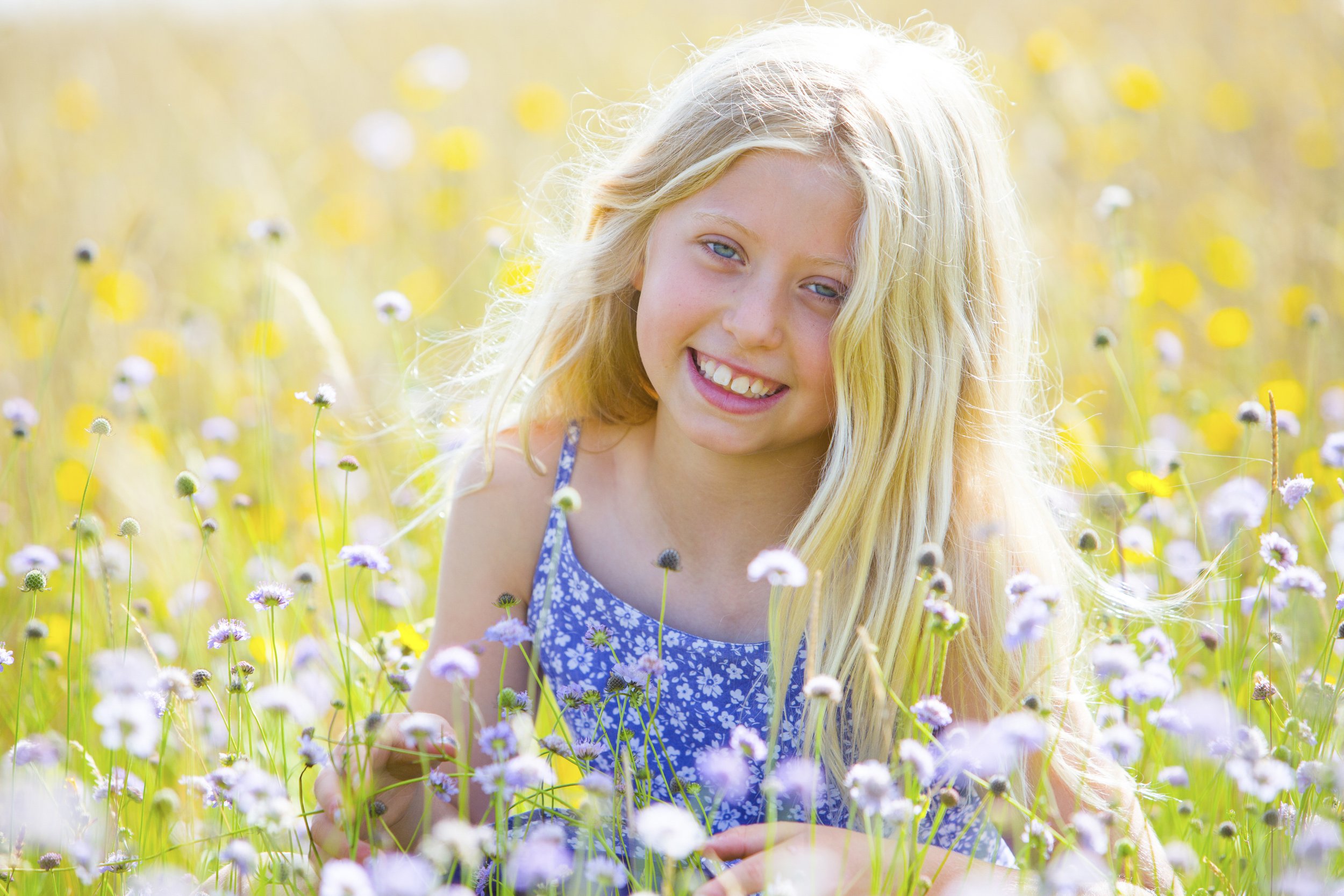A young girl with long blonde hair wearing a blue floral dress, smiling while lying in a field of purple and yellow flowers.