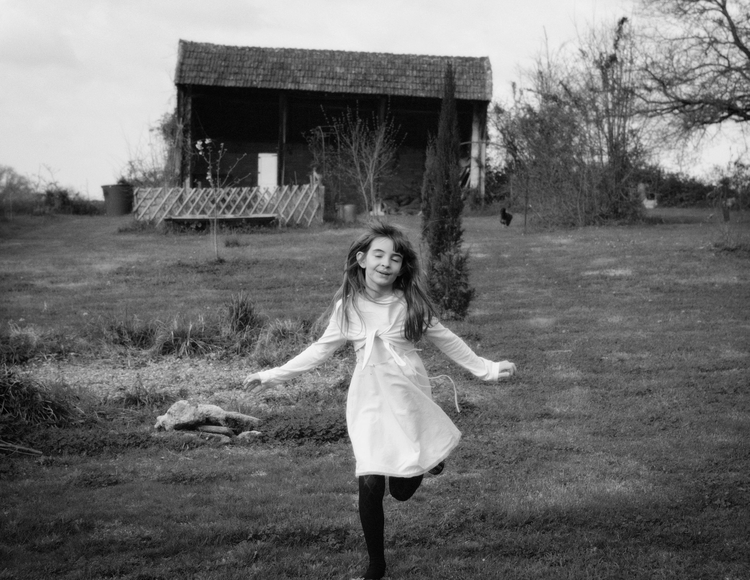 A girl playing outside in a yard with a large barn-like structure in the background, surrounded by trees and grass.