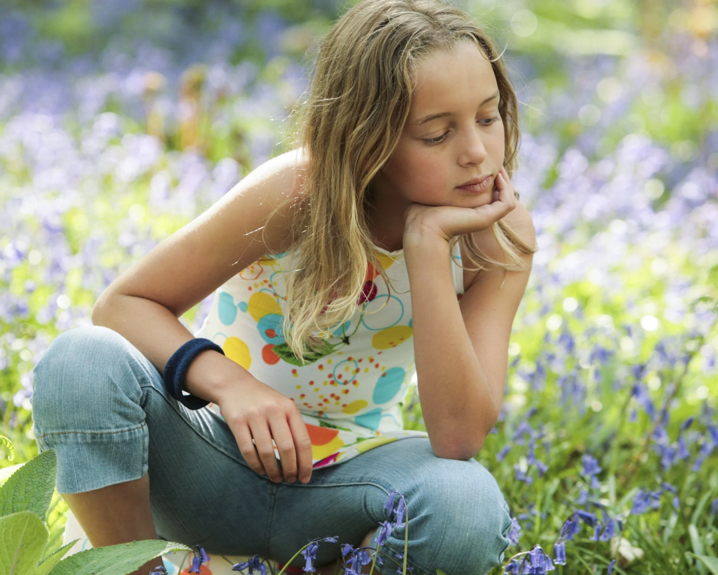 A young girl sitting in a field of blue and purple flowers, resting her chin on her hand and looking thoughtful.