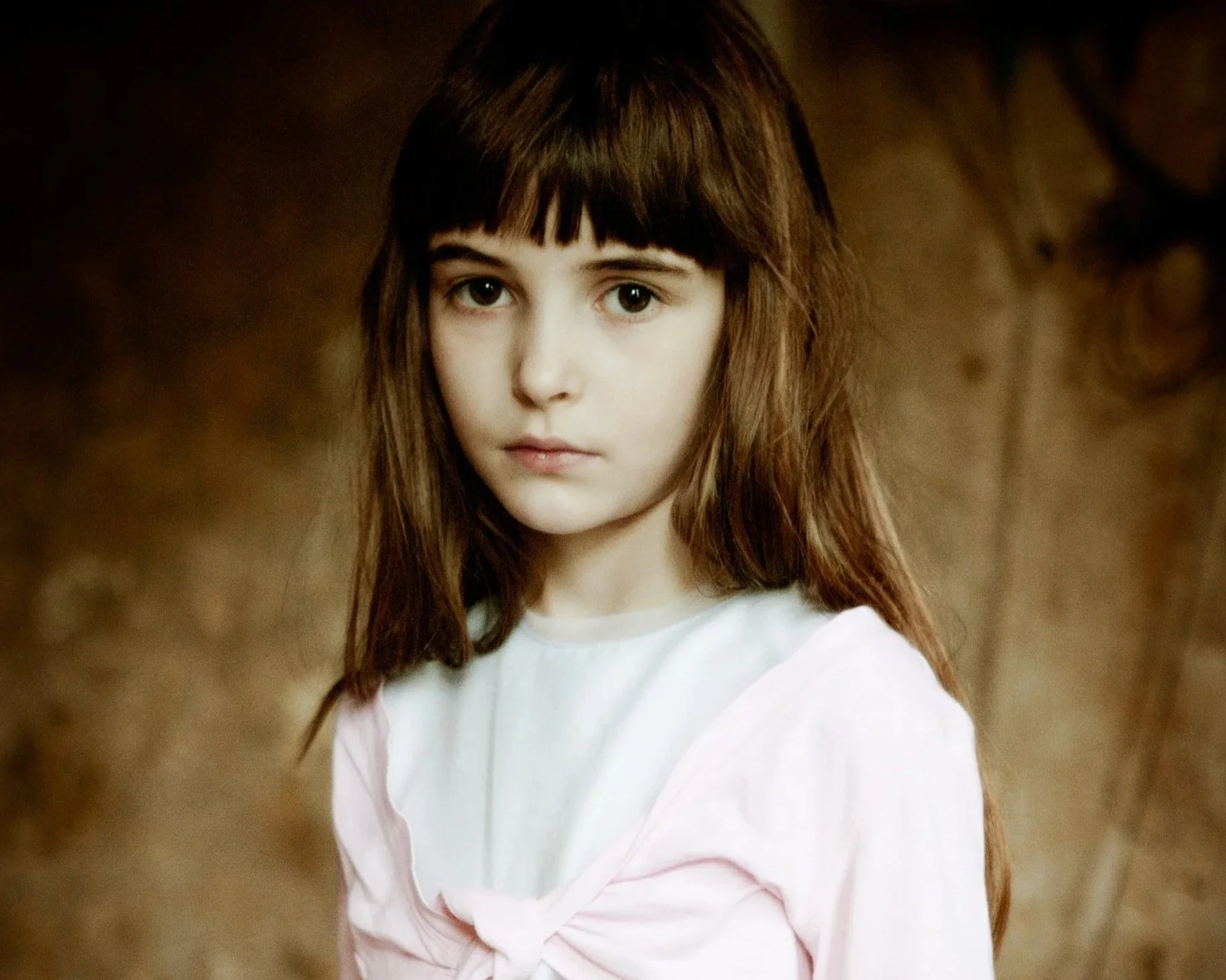 A young girl with long brown hair and bangs, wearing a light pink top, looking directly at the camera with a neutral expression, against a blurred brown background.