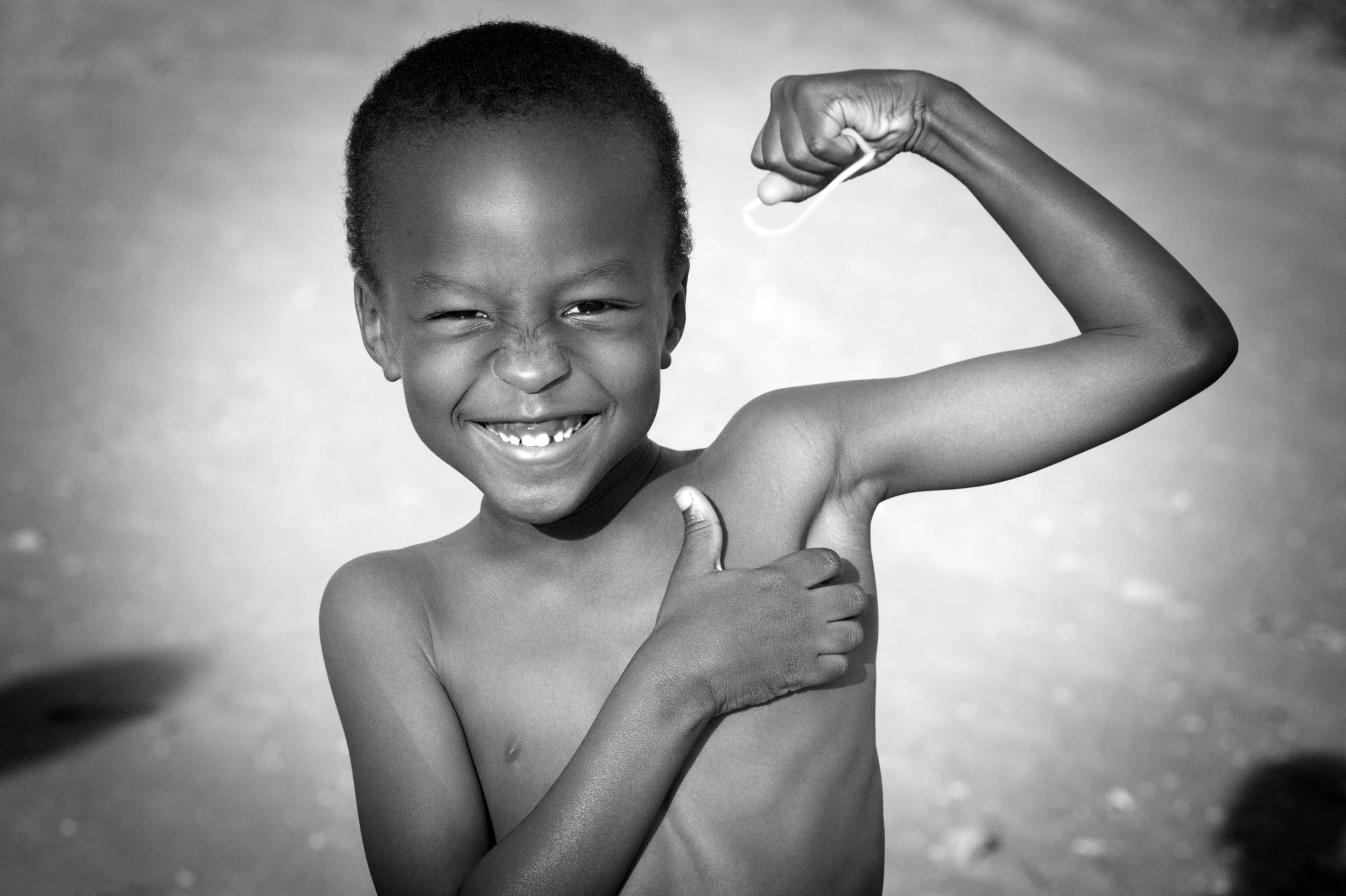 A young boy smiling and flexing his arm to show his bicep, holding a small object in his hand. The photo is in black and white.