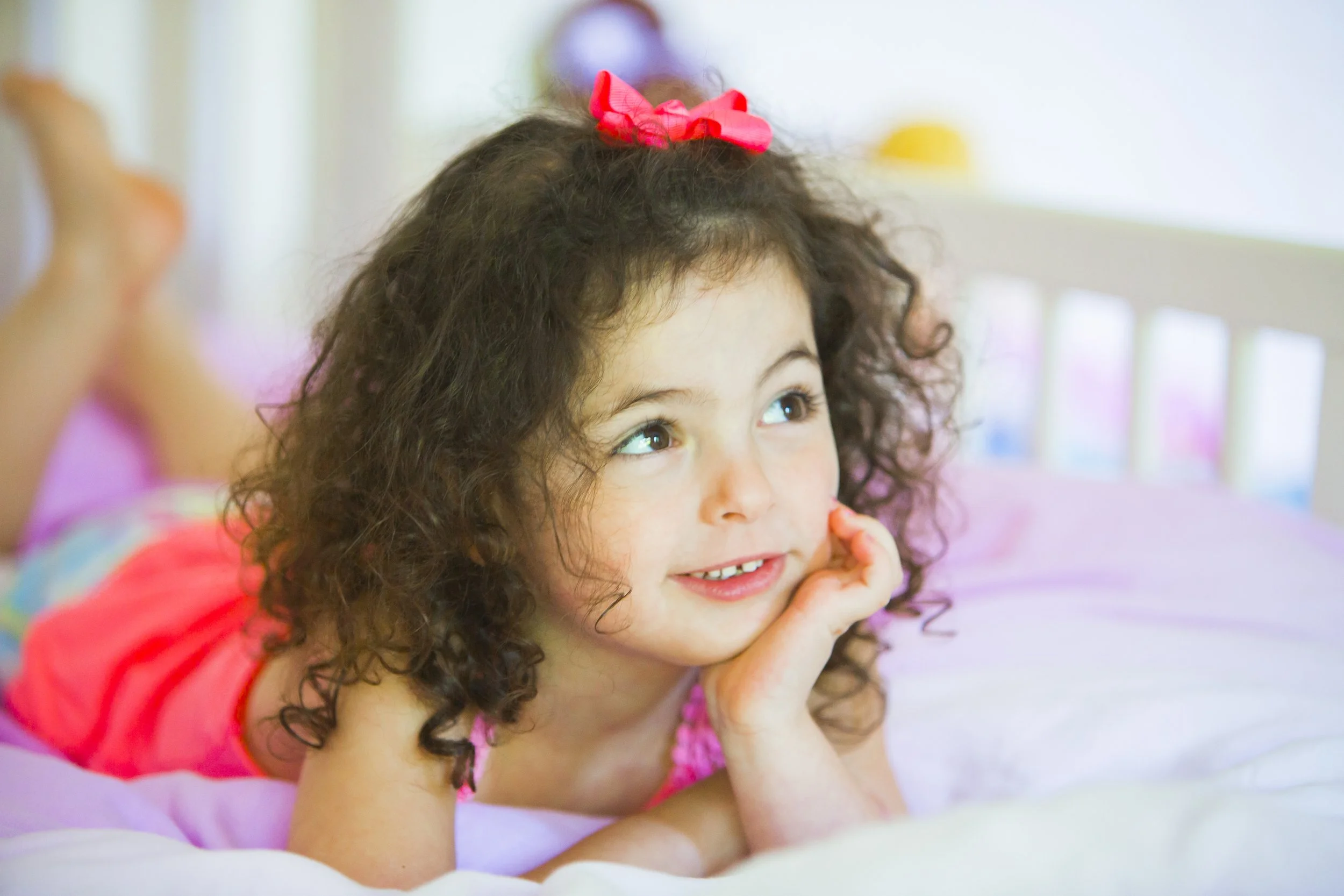 A young girl with curly hair lying on her stomach on a bed with her chin resting on her hands, smiling and looking to the side.