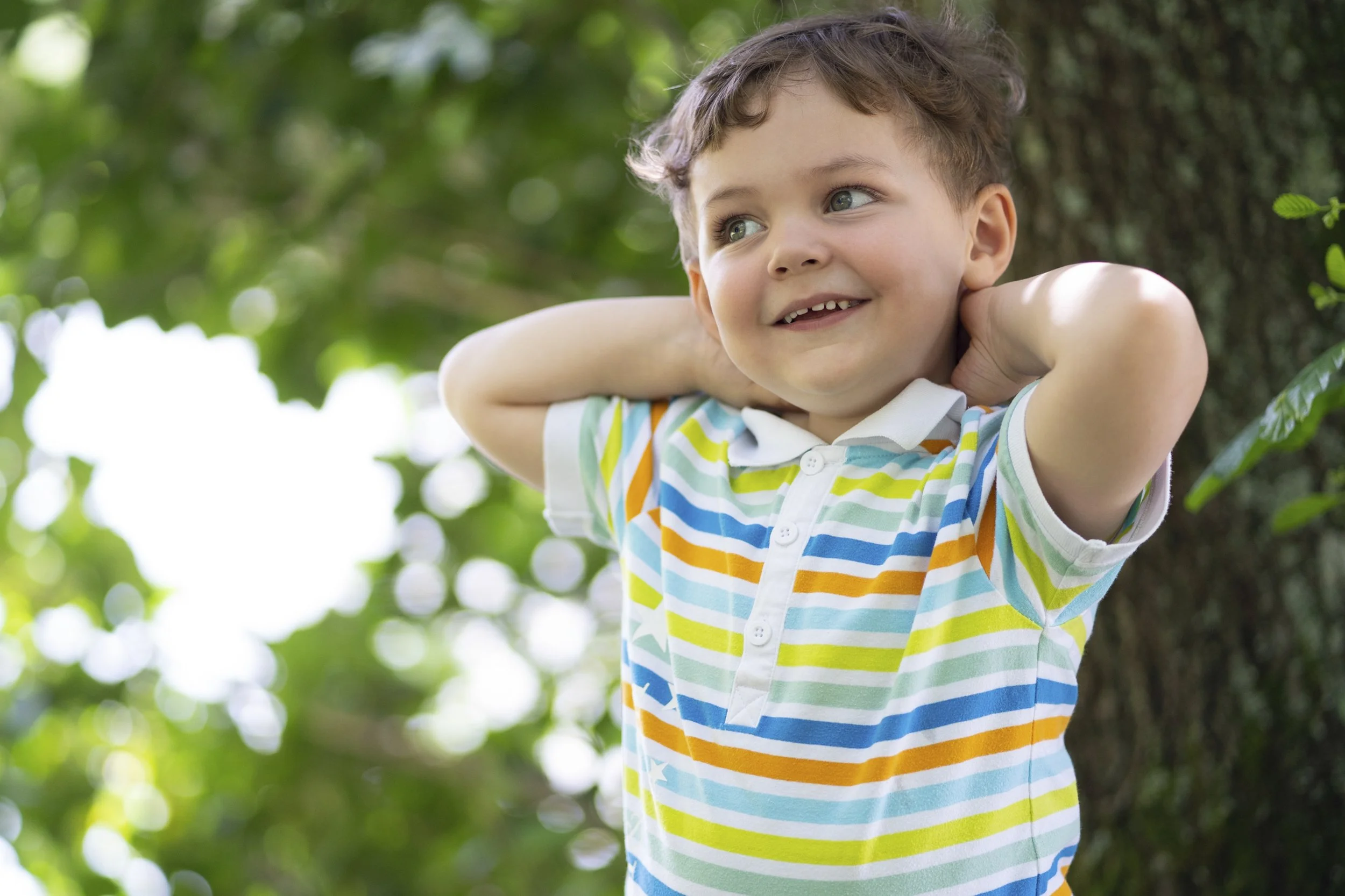 Young boy with curly brown hair smiling, standing outdoors with green leafy background, wearing a white polo shirt with colorful horizontal stripes, hands behind his head.