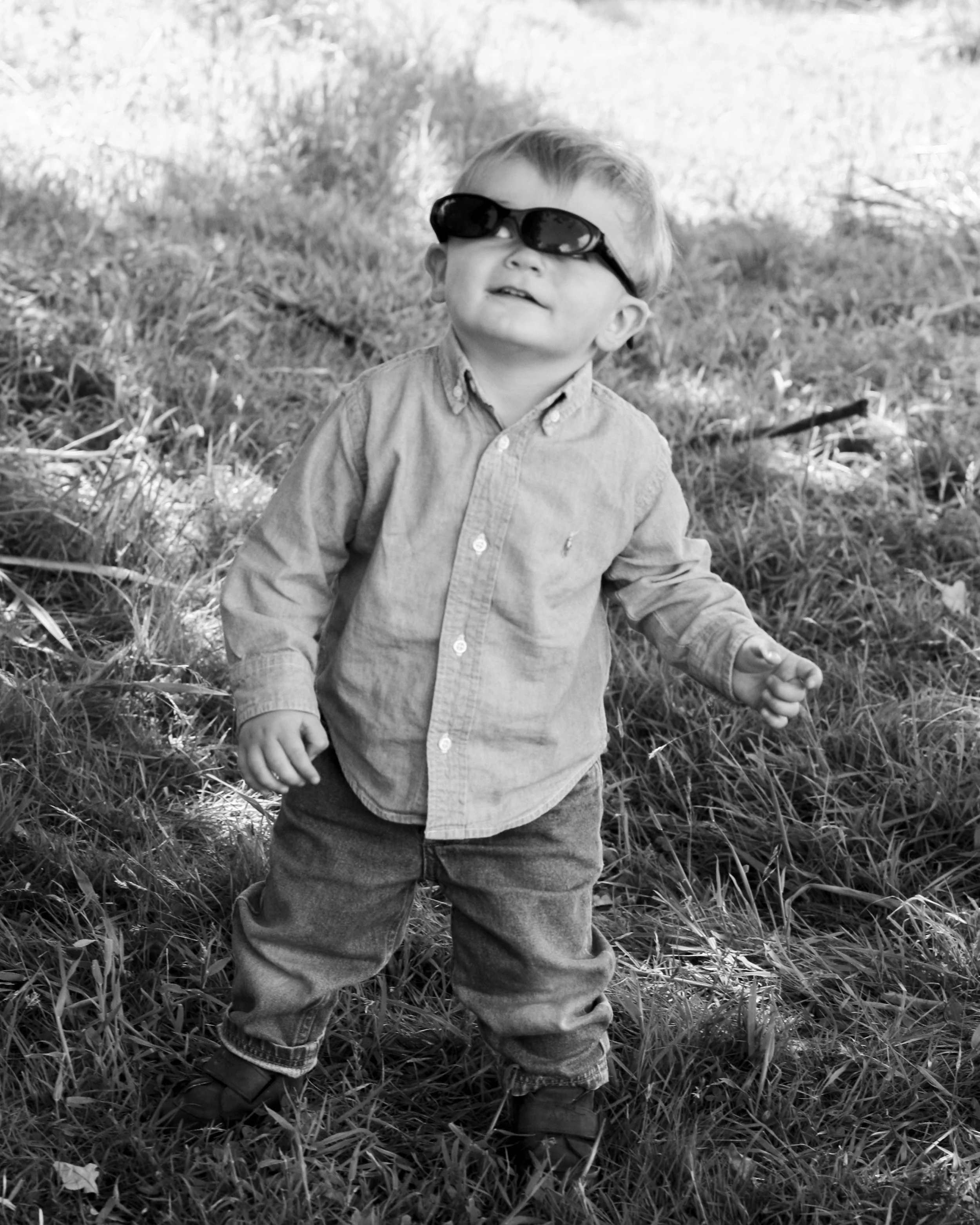 A young boy wearing sunglasses, a button-up shirt, jeans, and sneakers standing outdoors among grass and trees.