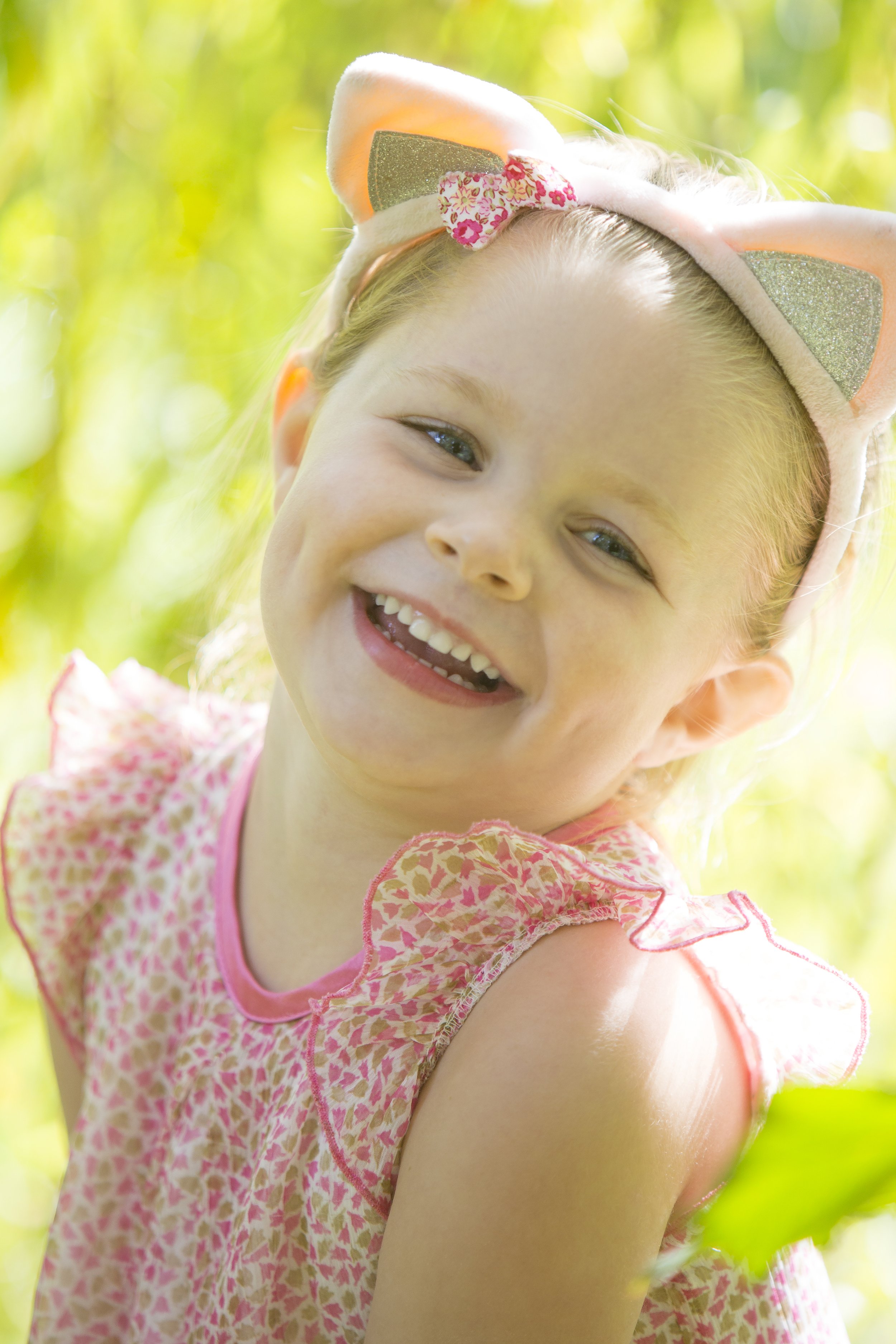 A young girl smiling and wearing a pink dress with ruffled sleeves and a headband with cat ears and pink floral bow, outdoors with greenery in the background.