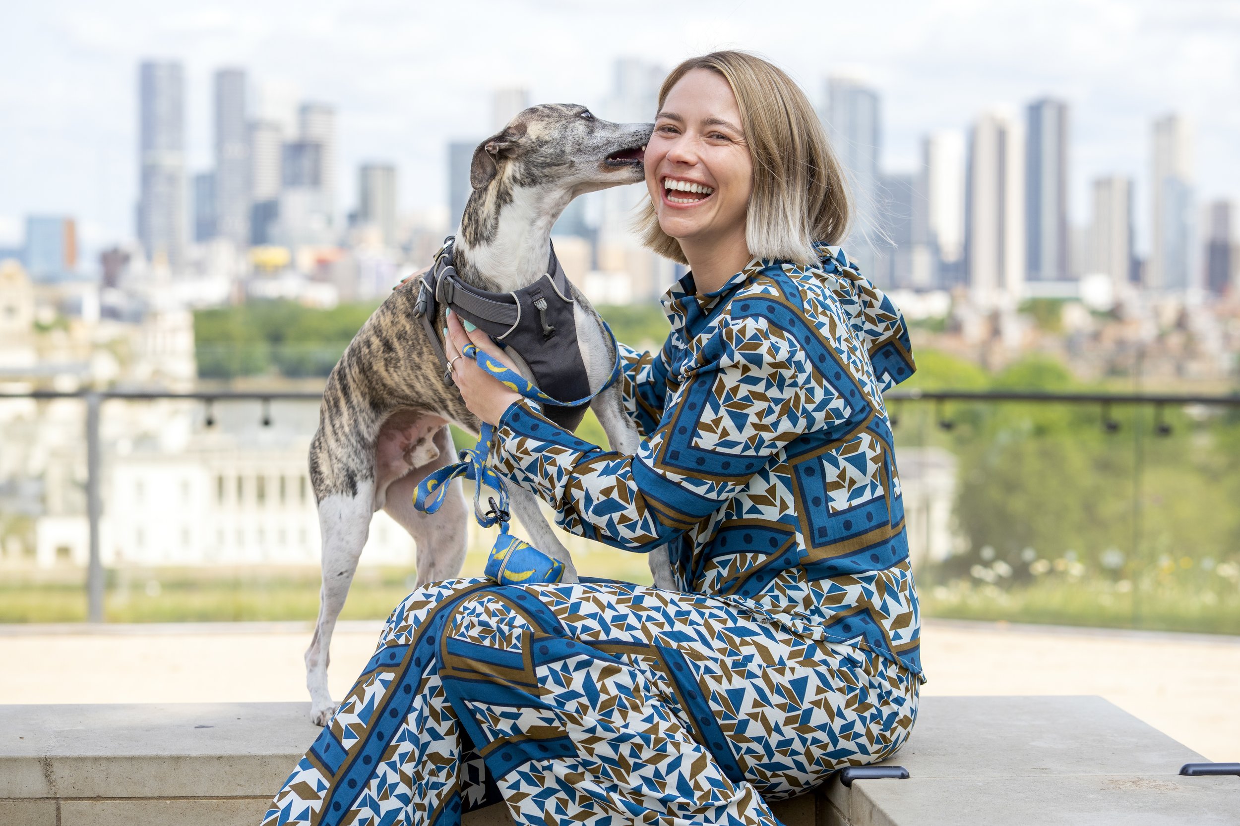 A woman with shoulder-length blonde hair is sitting on a concrete ledge, smiling as her dog licks her face. The dog is wearing a harness. The background shows a city skyline with tall buildings and a clear sky.