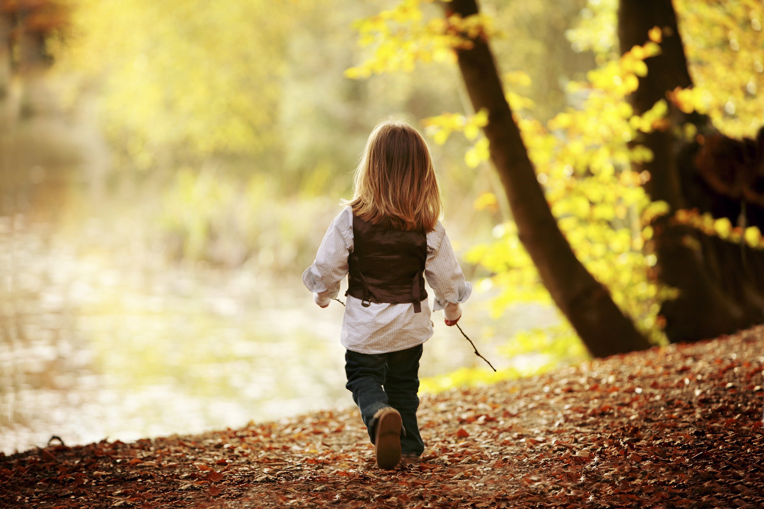 A young girl with brown hair walking through a forested area during autumn, holding a stick, with leaves on the ground and trees with yellow leaves in the background.