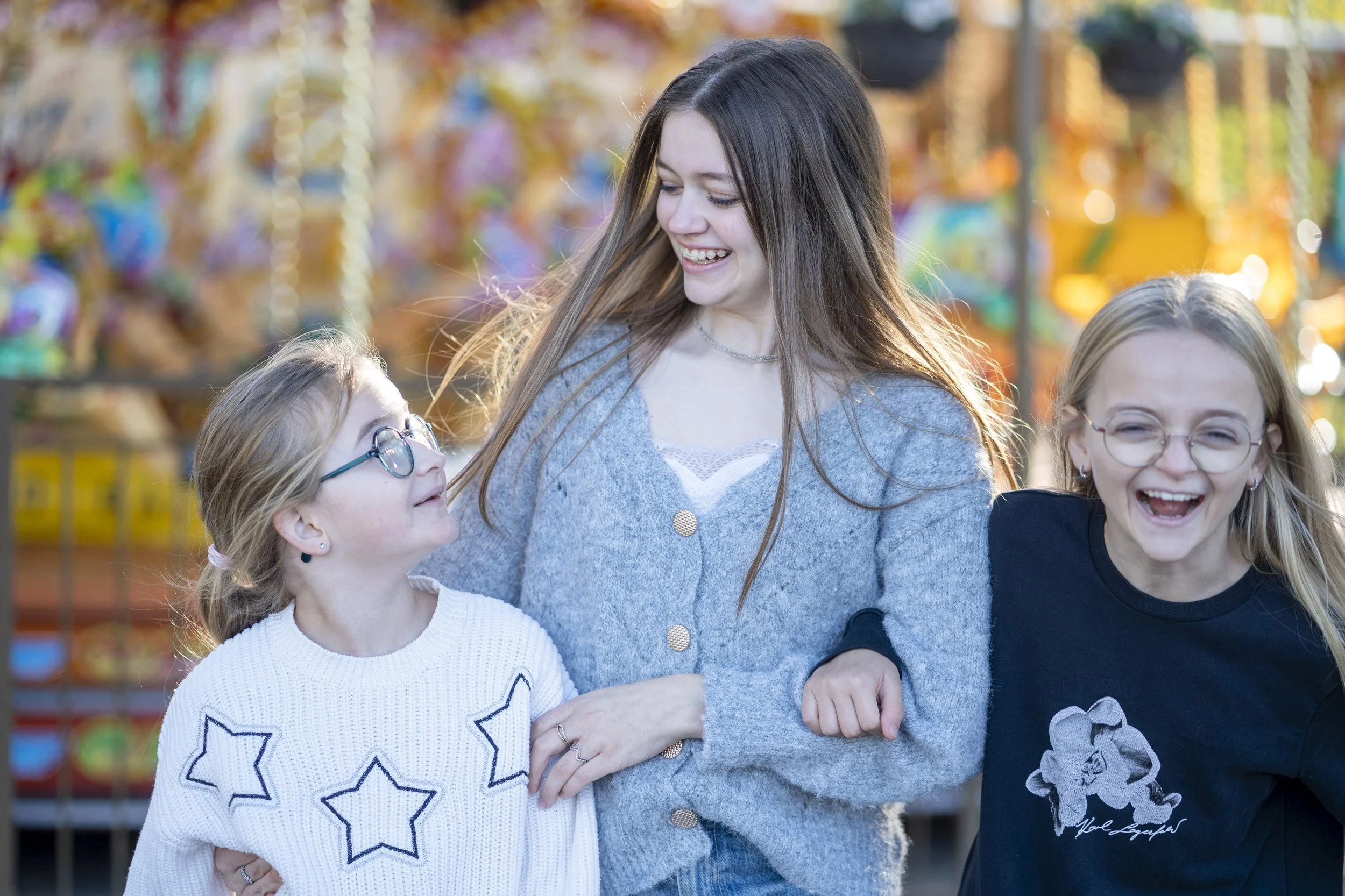 Three people, a woman and two girls, are smiling and enjoying a day at the amusement park. The background shows a colorful carousel.