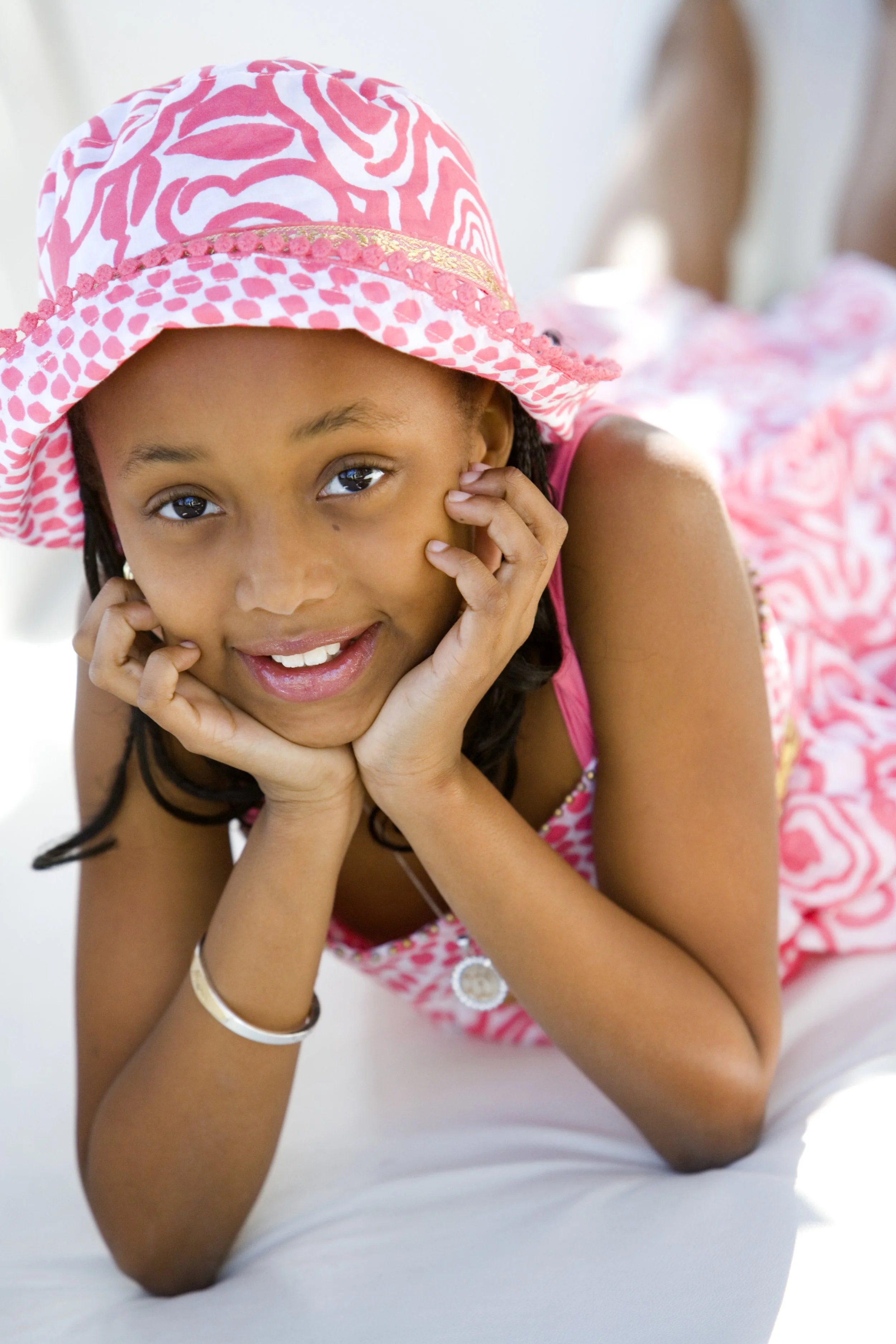 A young girl wearing a pink and white patterned hat and dress, lying on her stomach and smiling at the camera.
