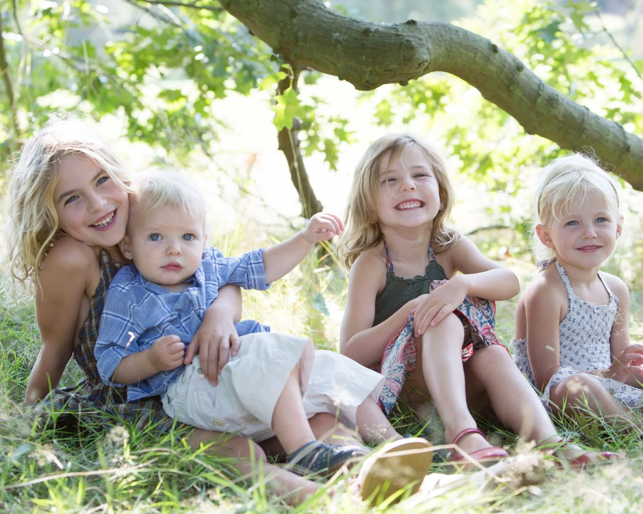 Four children, three girls and a boy, sitting on grass under a tree in a forest, smiling and enjoying a sunny day.