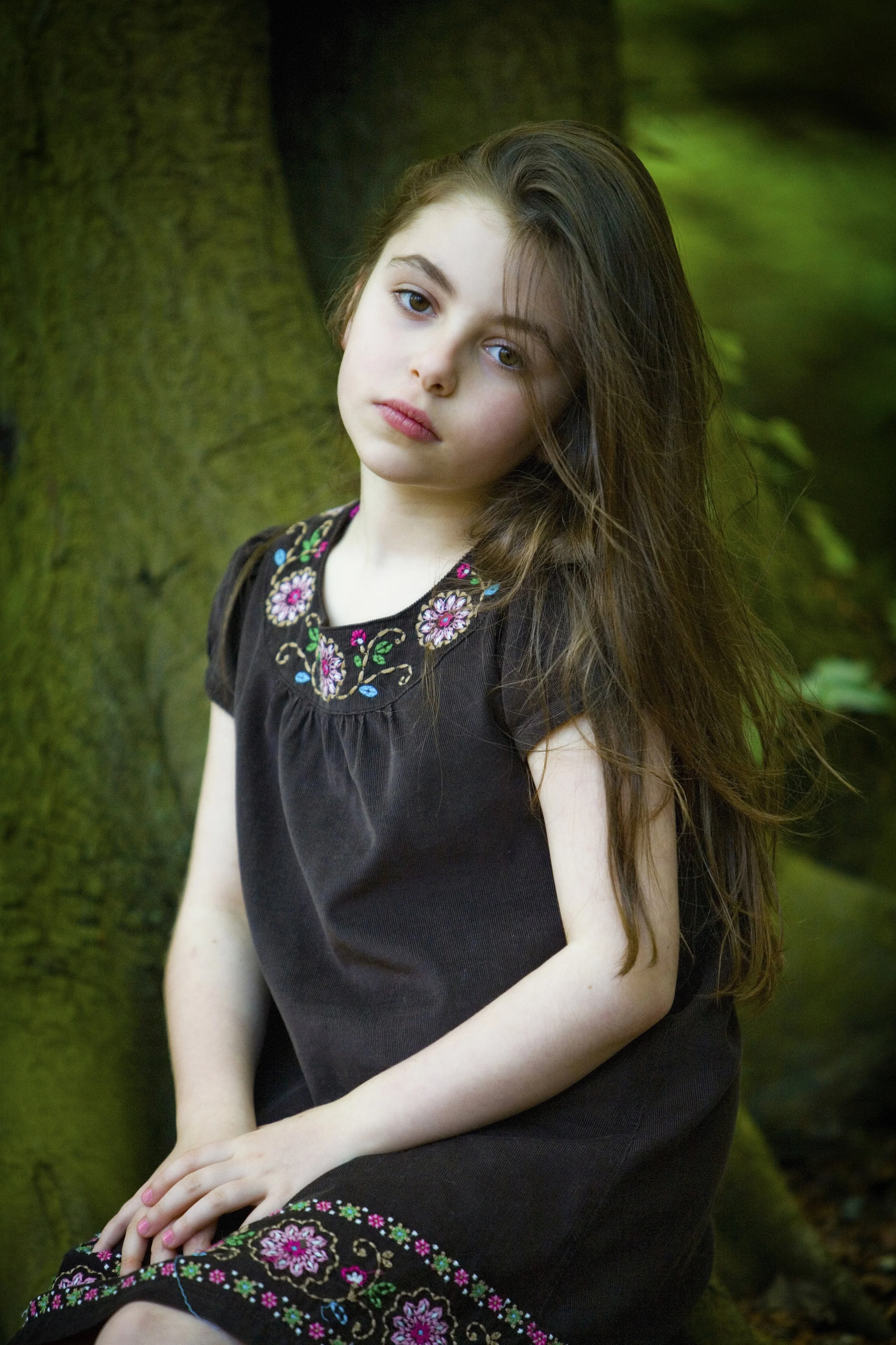 A young girl with long brown hair and fair skin sitting outdoors near a tree, wearing a black dress with colorful floral embroidery around the neckline and hem.