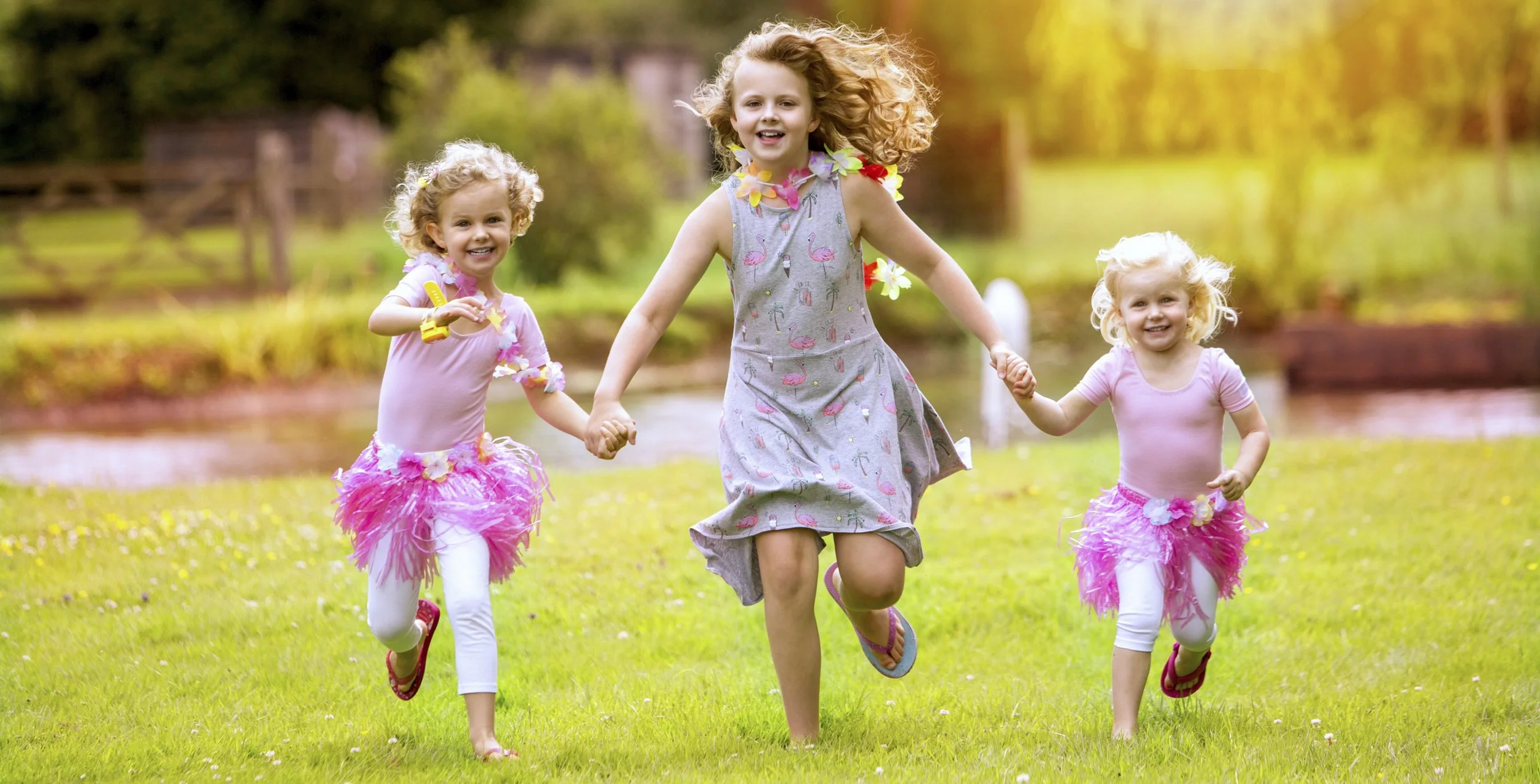 Three young girls running and playing outdoors on green grass, holding hands. They are wearing colorful outfits with pink feathered skirts and leis around their necks, enjoying a sunny day in a park or garden.