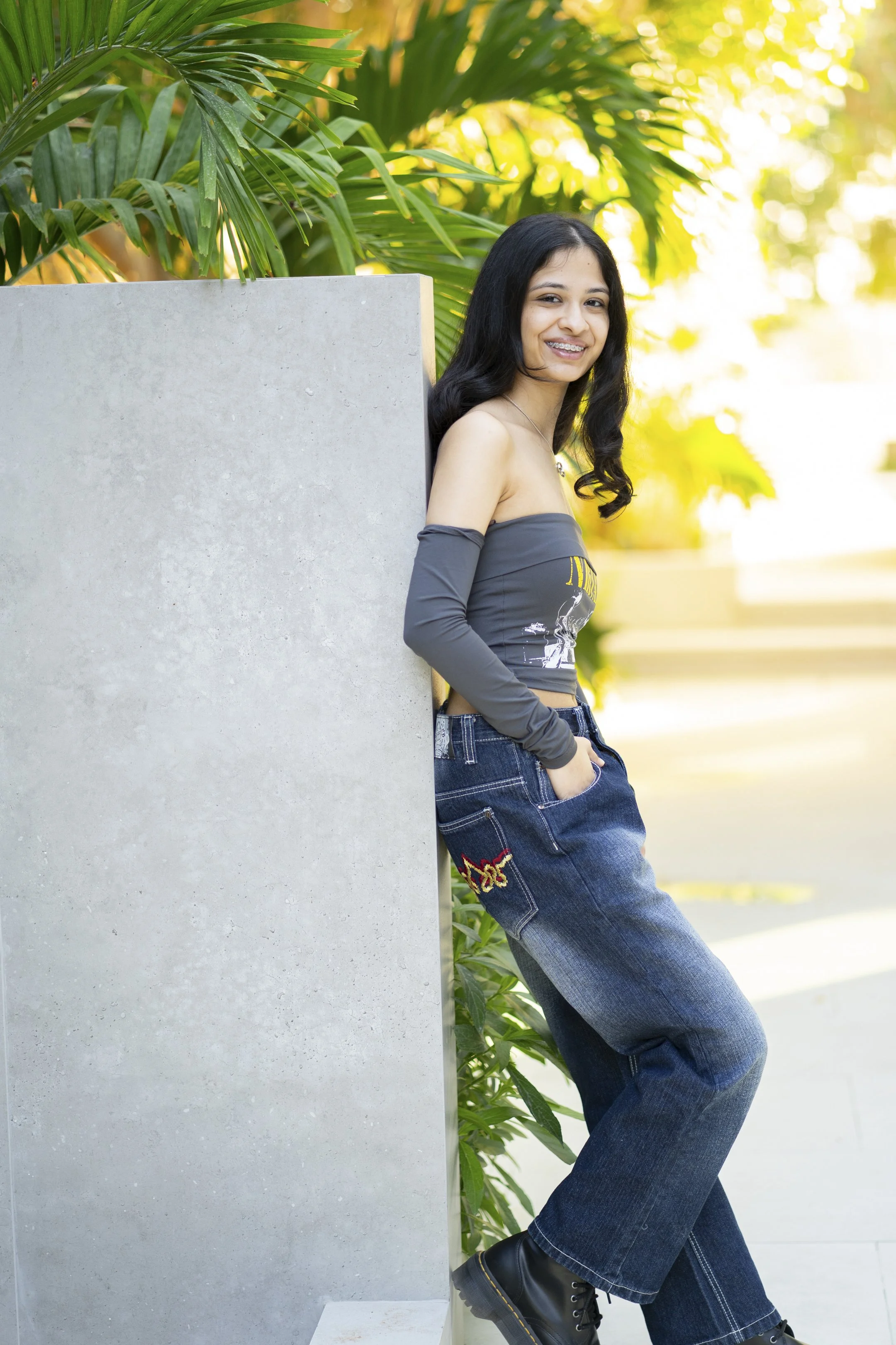 A young woman with long black hair, smiling and leaning against a concrete wall outdoors. She is wearing a gray off-the-shoulder top, wide-leg jeans with embroidered details, and black boots, surrounded by green tropical plants with sunlight filterin