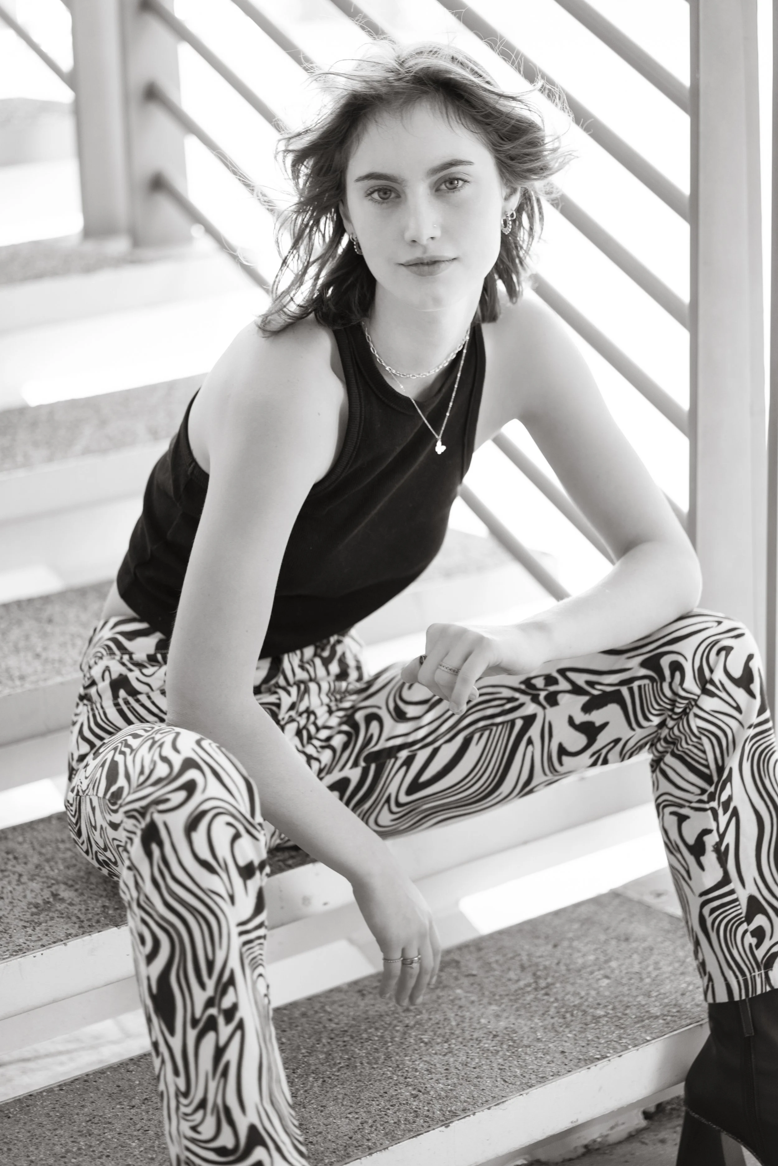 A young woman with shoulder-length wavy hair, wearing a sleeveless black top, patterned zebra-style pants, and accessories including necklaces, earrings, and rings, sitting on outdoor stairs.
