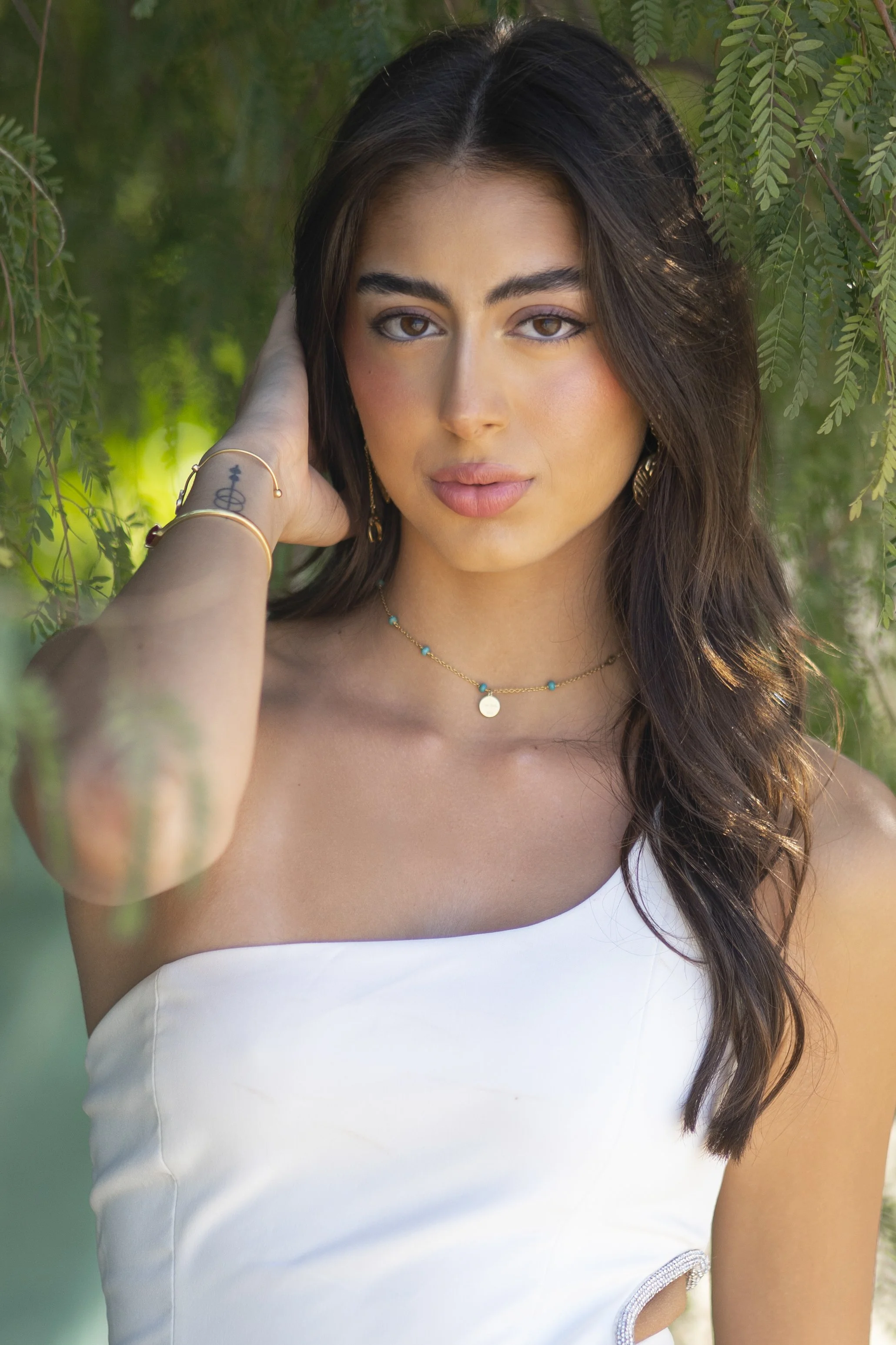 A woman with dark brown hair and light brown eyes is outdoors, wearing a white strapless top and gold jewelry, with green foliage in the background.
