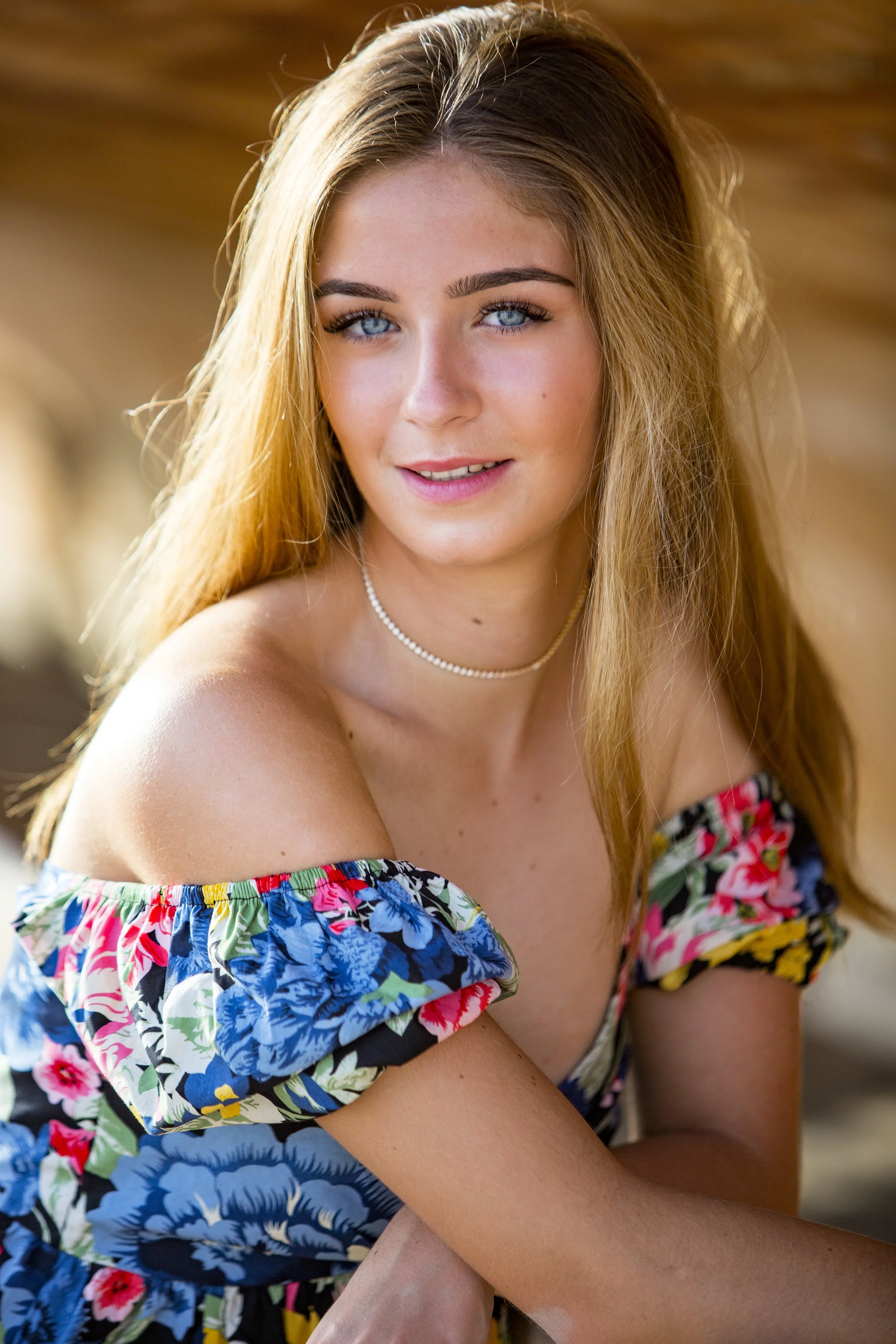 A young woman with long, light brown hair and blue eyes, wearing an off-the-shoulder floral dress and a pearl necklace, smiling outdoors.