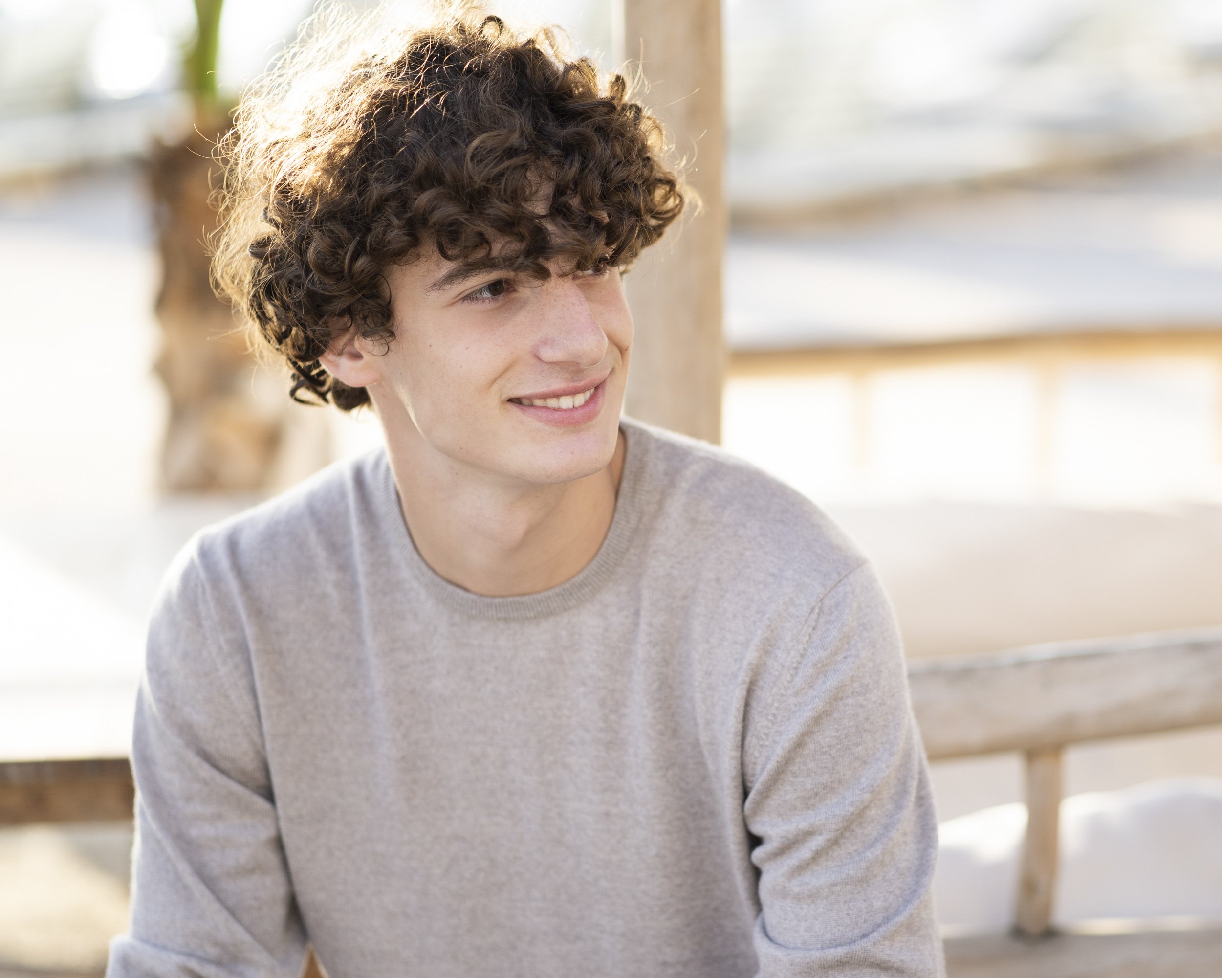 A young man with curly brown hair, wearing a light gray sweater, smiling and looking to his right, sitting outdoors near a wooden structure with blurred background.
