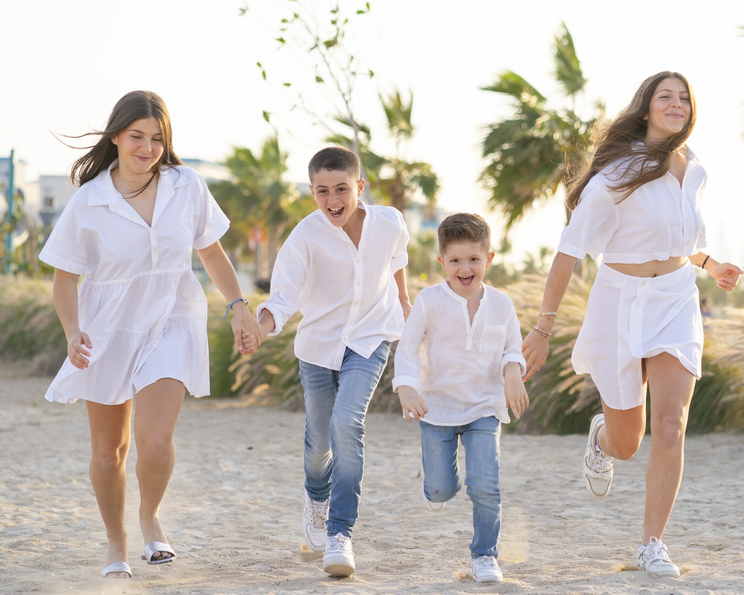 A family of four happily running on a sandy beach with palm trees in the background, dressed in white clothes.