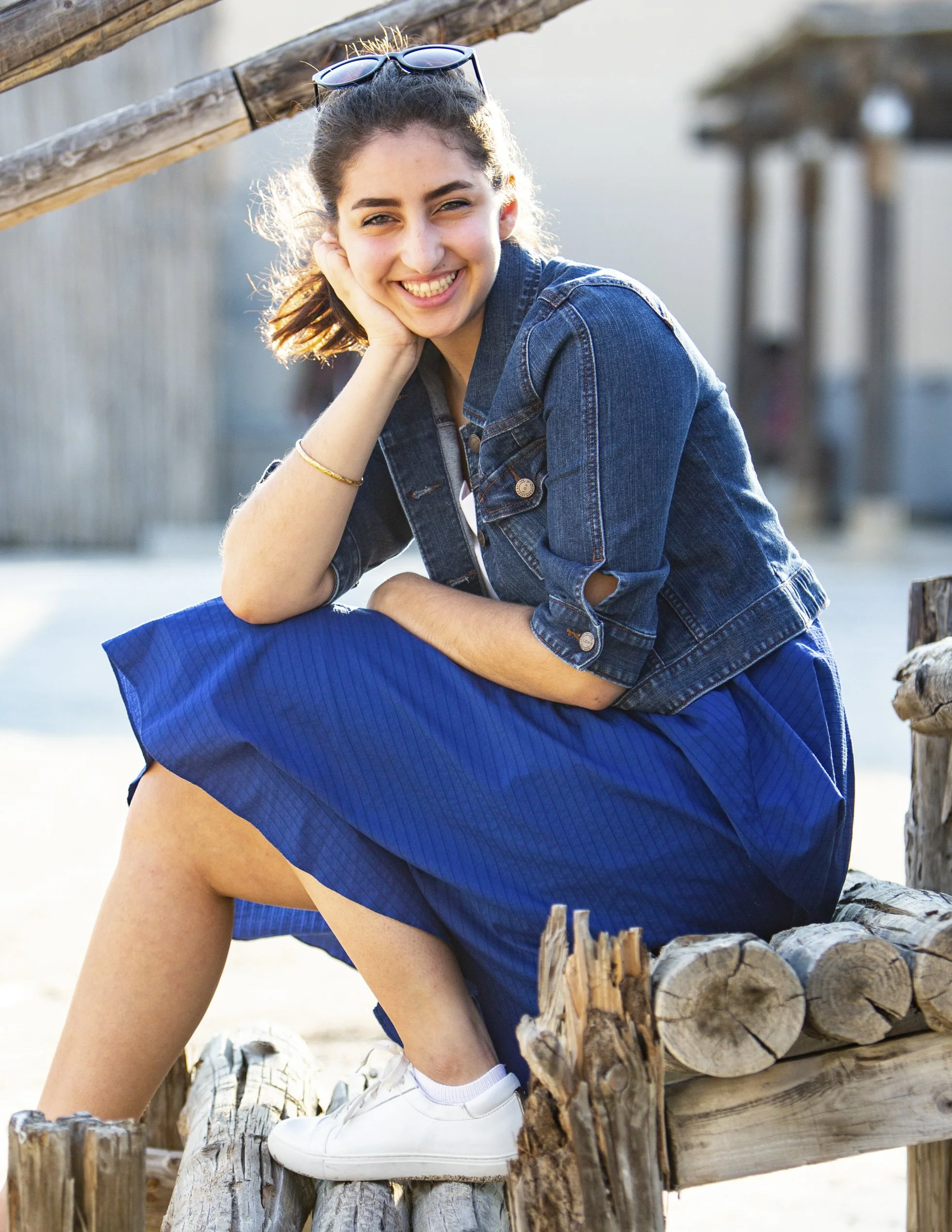 Young woman sitting outdoors on a wooden bench, smiling at the camera, wearing a denim jacket, blue skirt, and white sneakers, with sunglasses on her head.