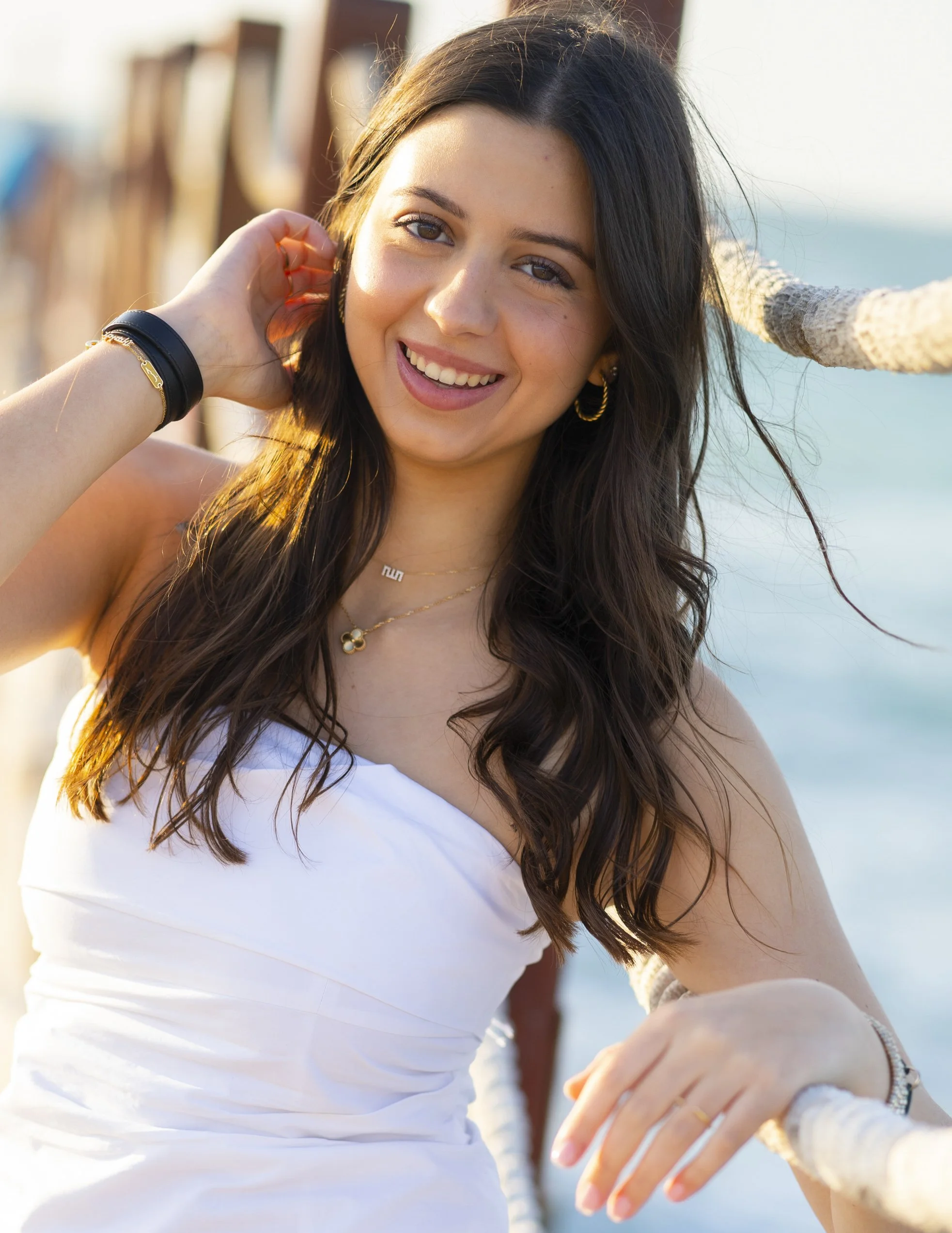 A young woman with long dark hair smiling, wearing a white dress, jewelry, and sunglasses, standing on a beach next to a rope railing.