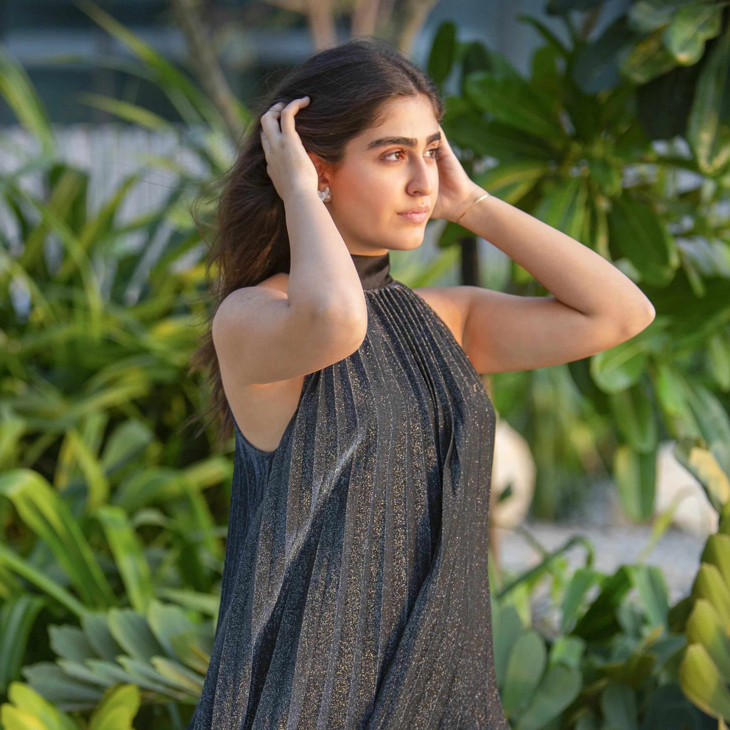 A young woman in a sleeveless, black, sparkly dress outdoors with lush green plants in the background, touchingt her hair and gazing off into the distance.