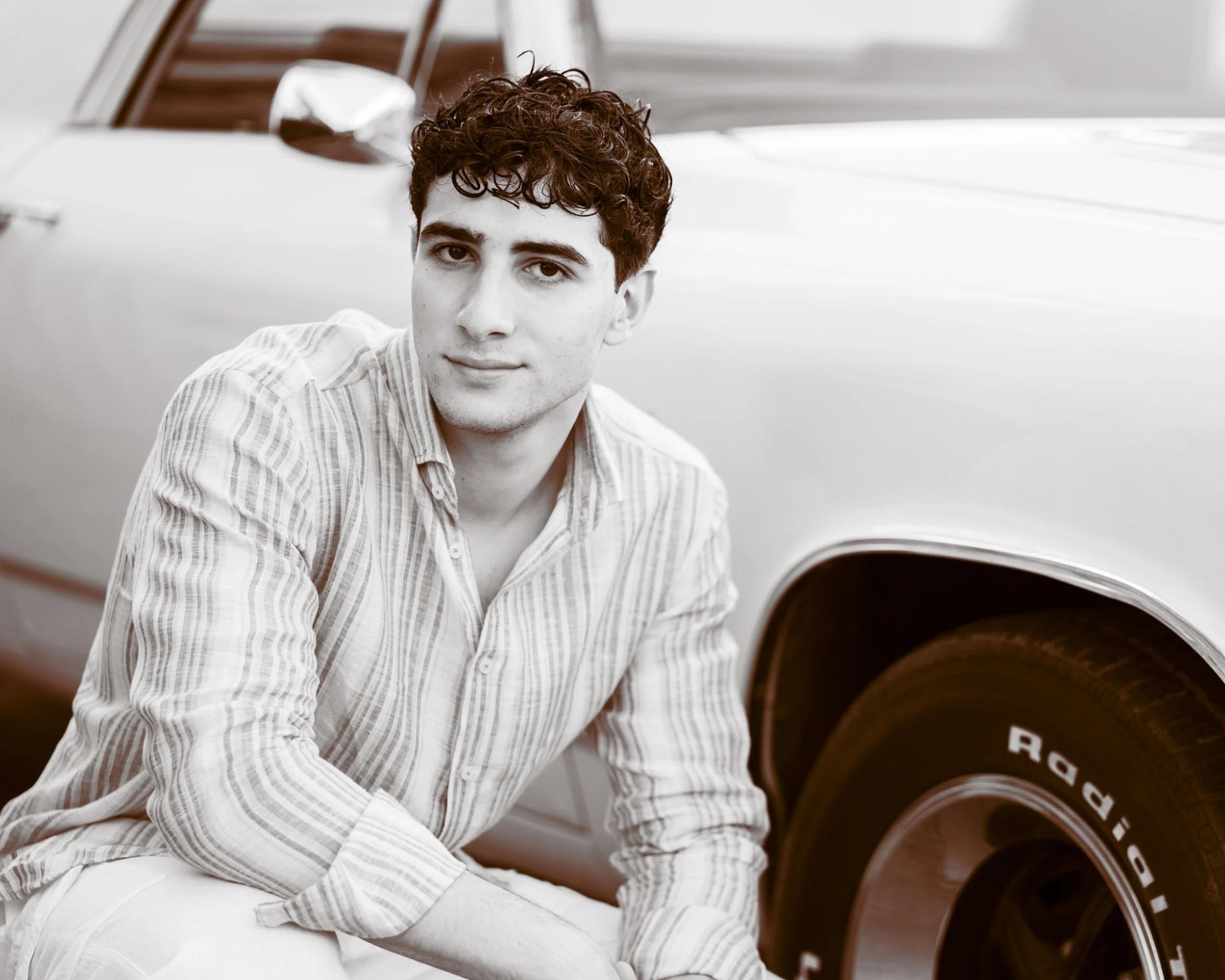 A young man with dark, curly hair wearing a striped shirt sitting next to a car tire.