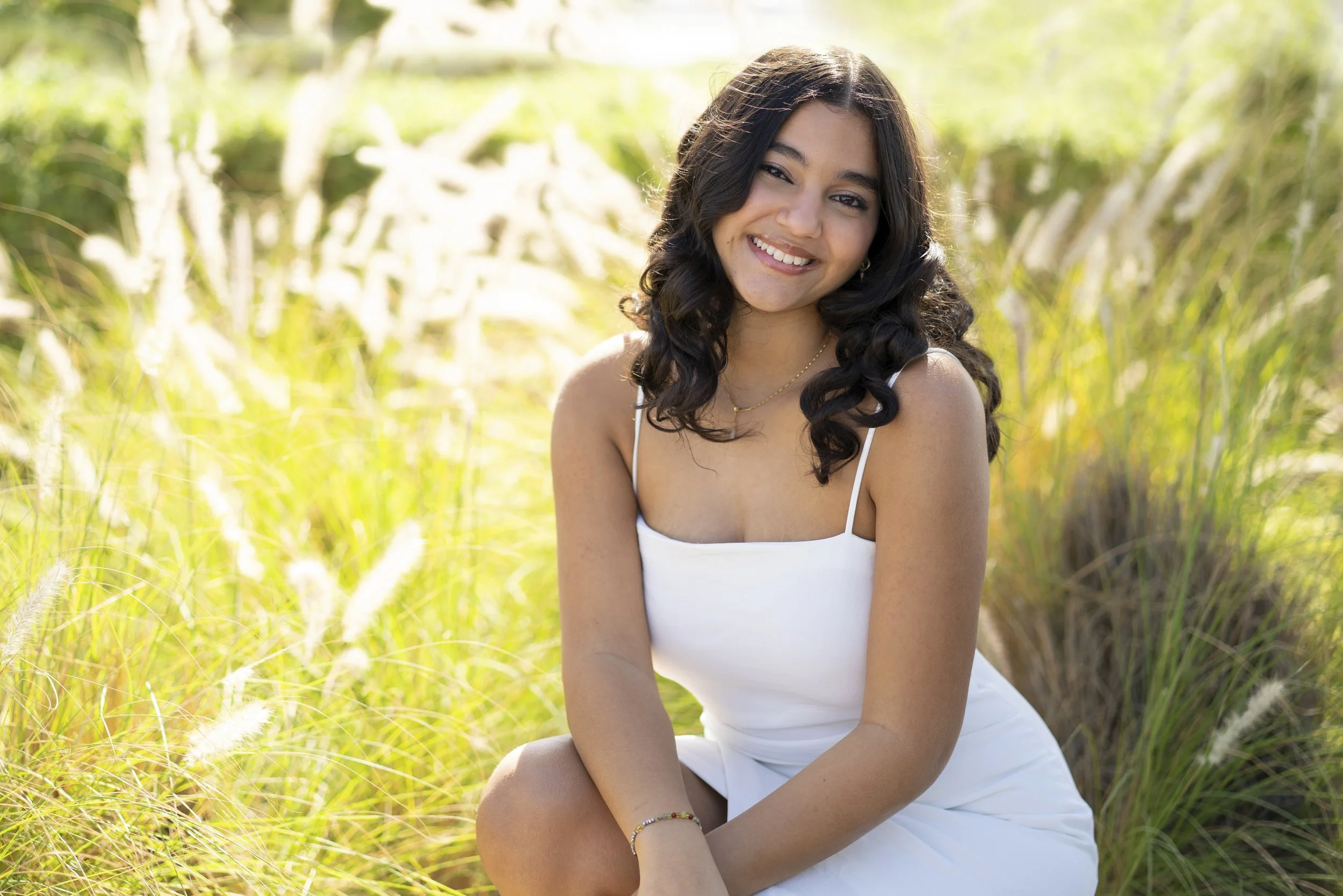 A young woman with dark, curly hair, wearing a white dress with thin straps, sitting outdoors in a grassy area, smiling at the camera.
