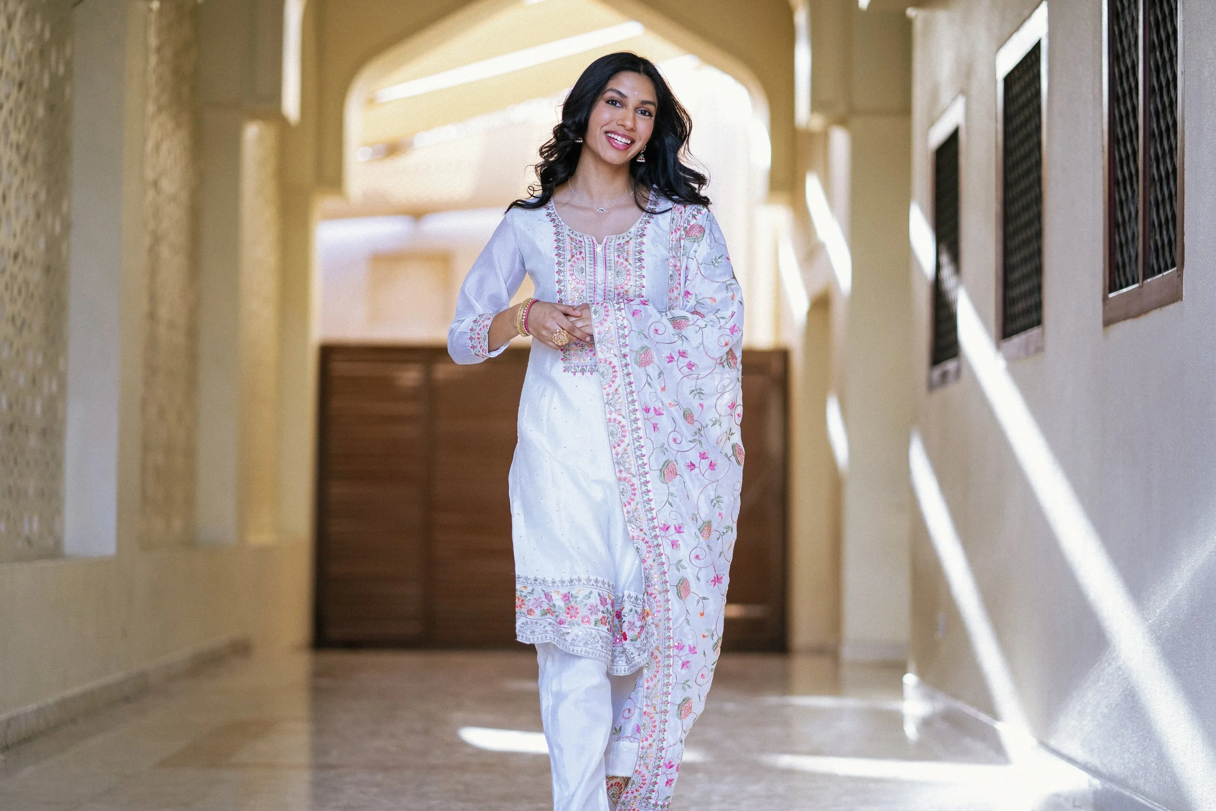 A woman dressed in a white embroidered traditional outfit walking down a brightly lit corridor, smiling and looking at the camera.