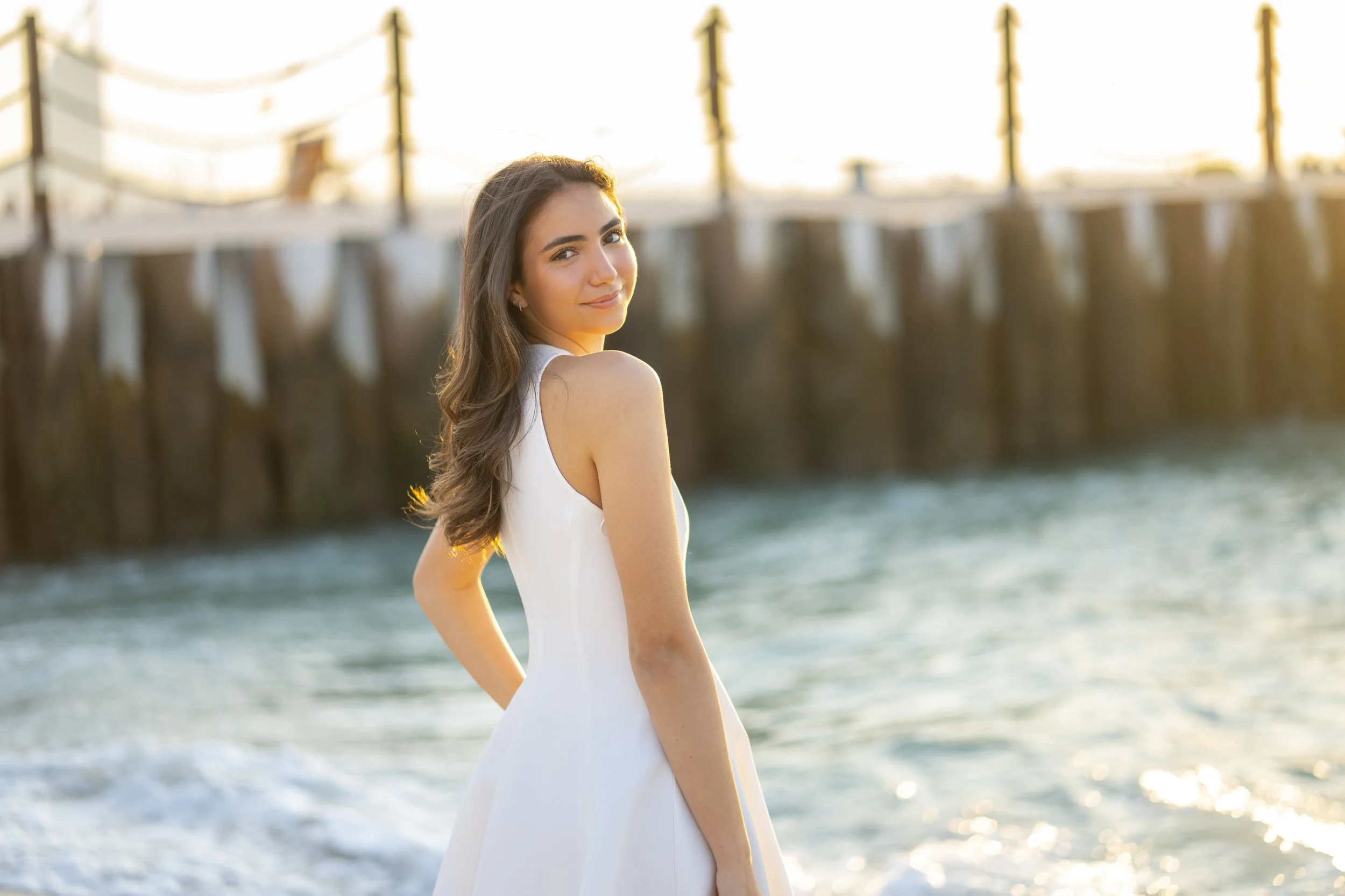 A young woman in a white dress standing on a beach near the water with a wooden pier in the background during sunset.