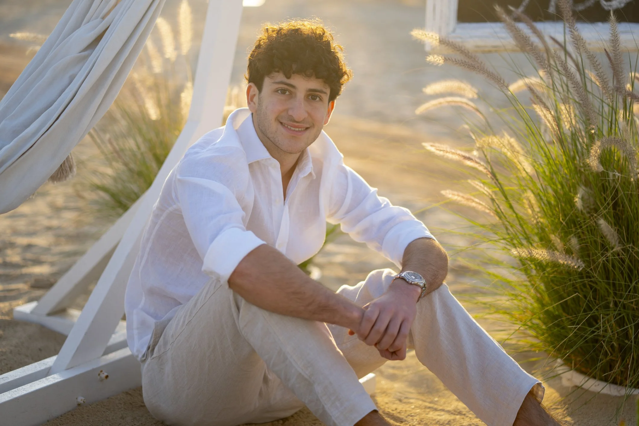 A smiling young man sitting on the sandy beach near grasses with a beach cabana in the background.