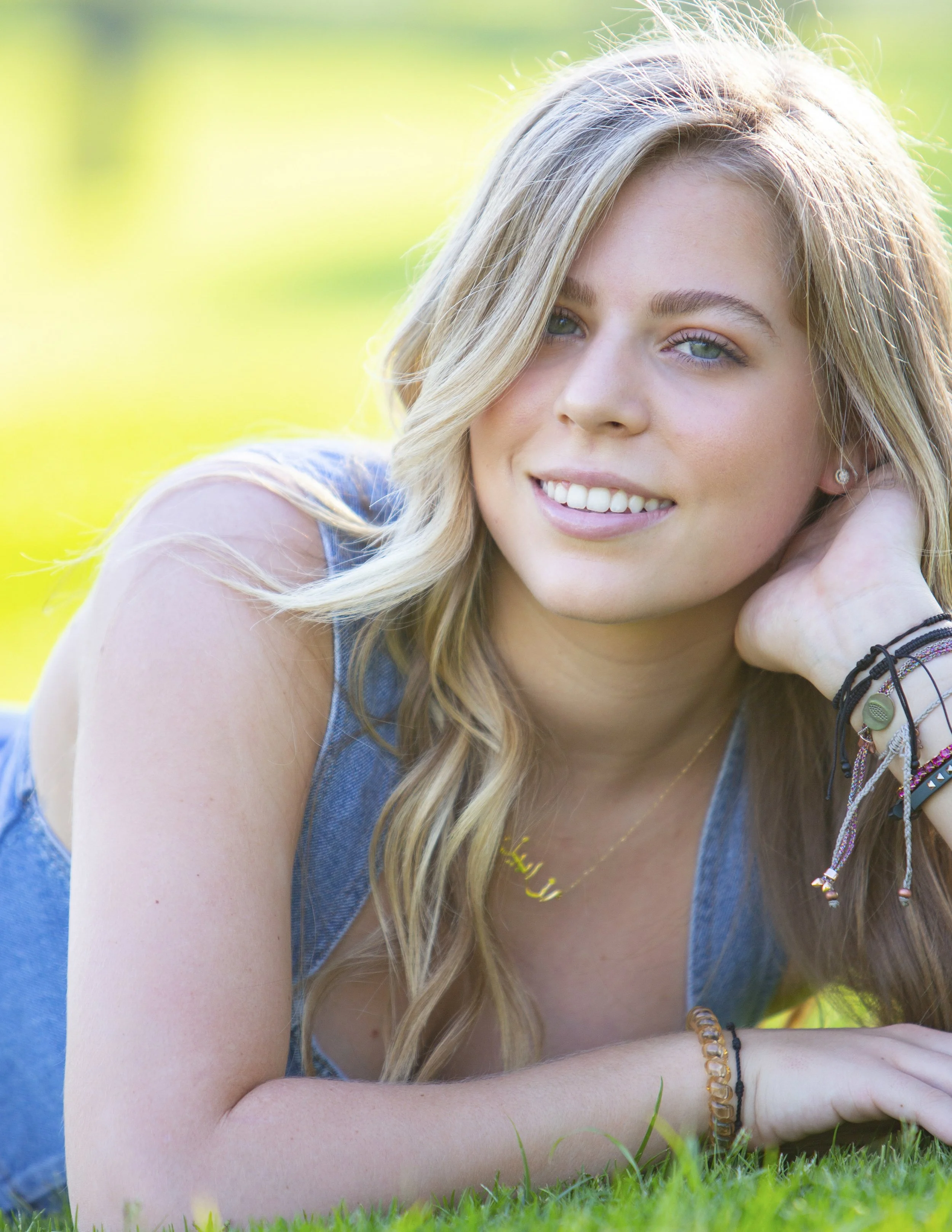 Young woman with wavy blonde hair lying on grass outdoors, smiling with blue eyes, wearing denim top and bracelets.