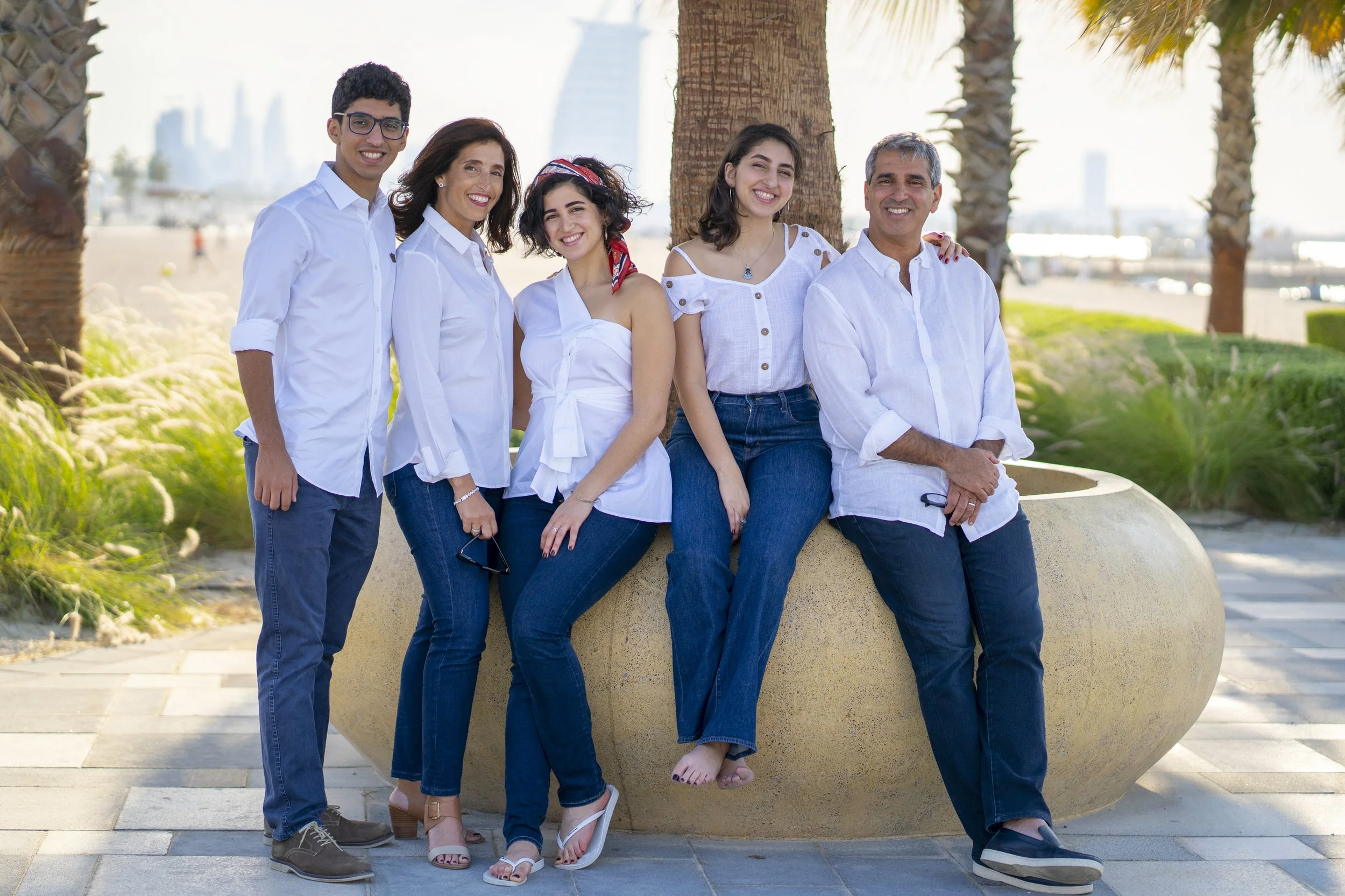 A family of six, dressed in white and blue, sitting and standing on a large round stone bench outdoors with palm trees and city skyline in the background.