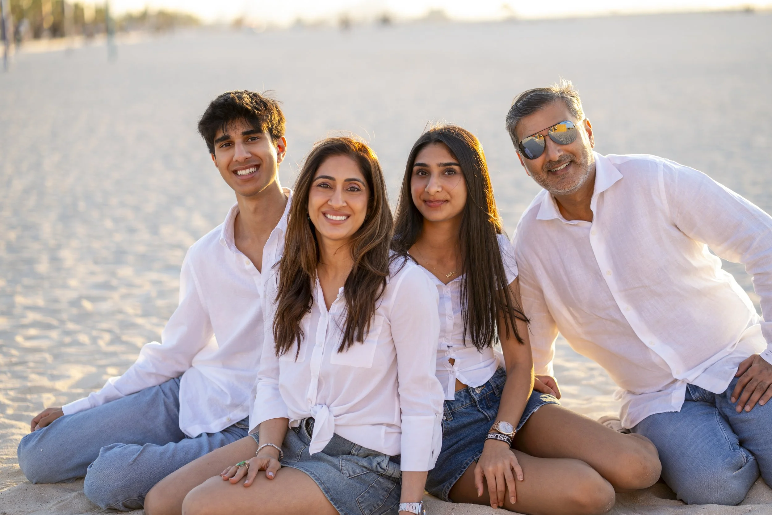Four people sitting on a sandy beach, enjoying the sunset, wearing white shirts and casual shorts.
