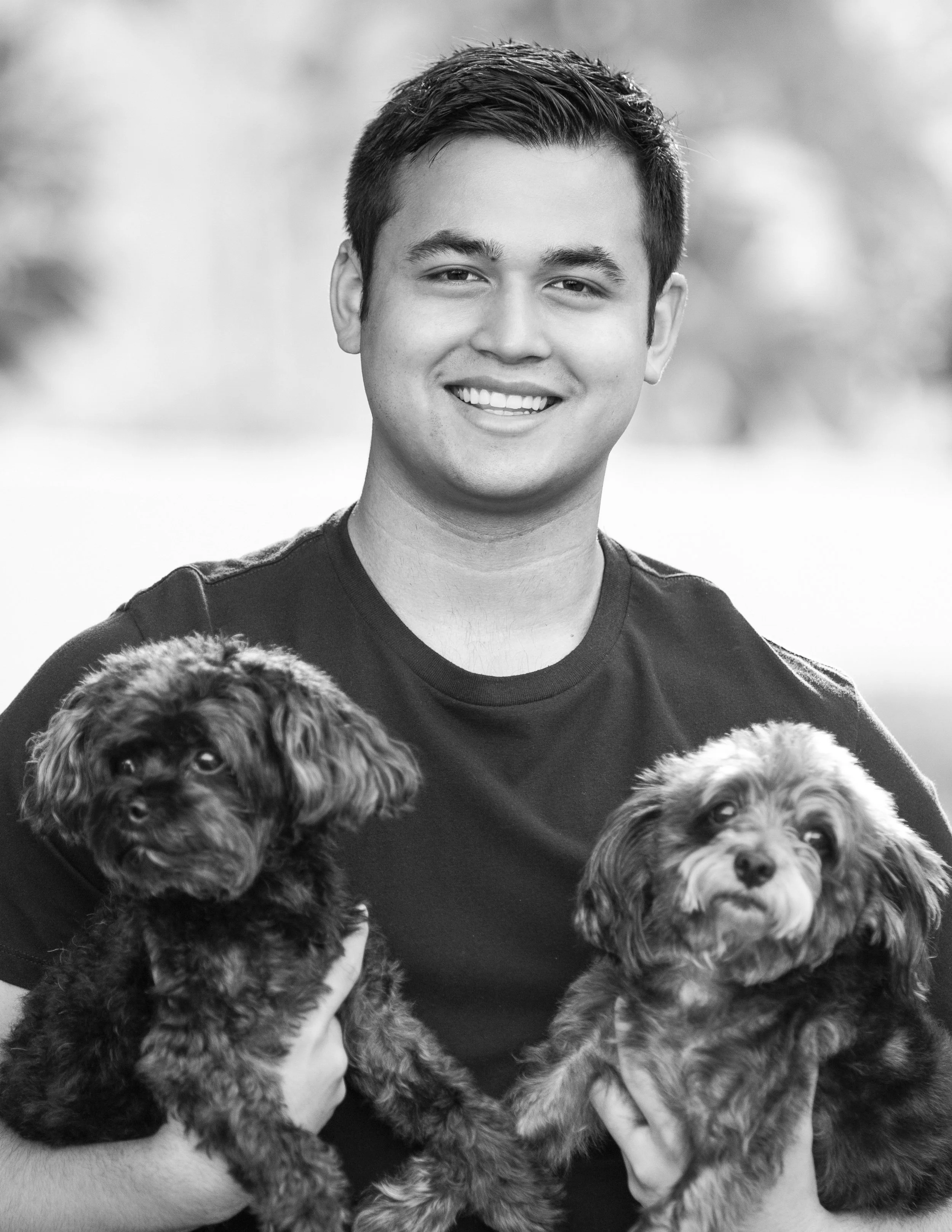 Black and white photo of a young man smiling, holding two small dogs, one in each hand, outdoors with blurred trees in the background.