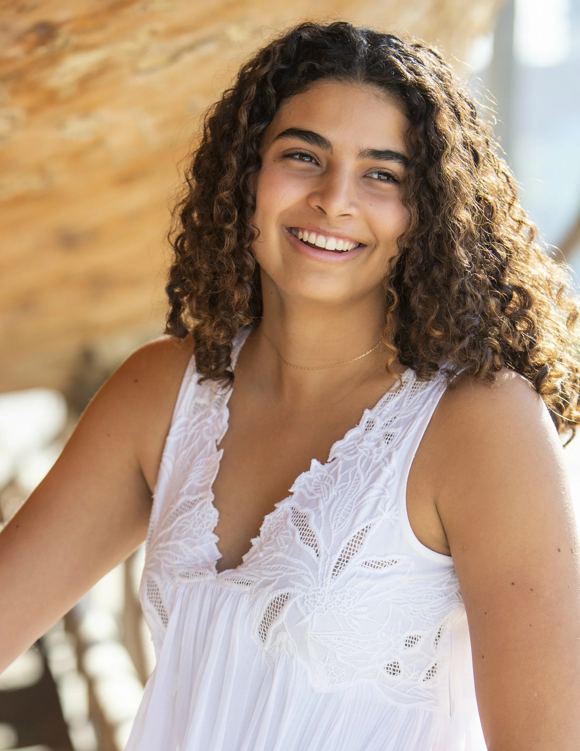 Portrait of a young woman with curly hair, smiling, wearing a white lace top, sitting indoors with a blurred brick wall background.
