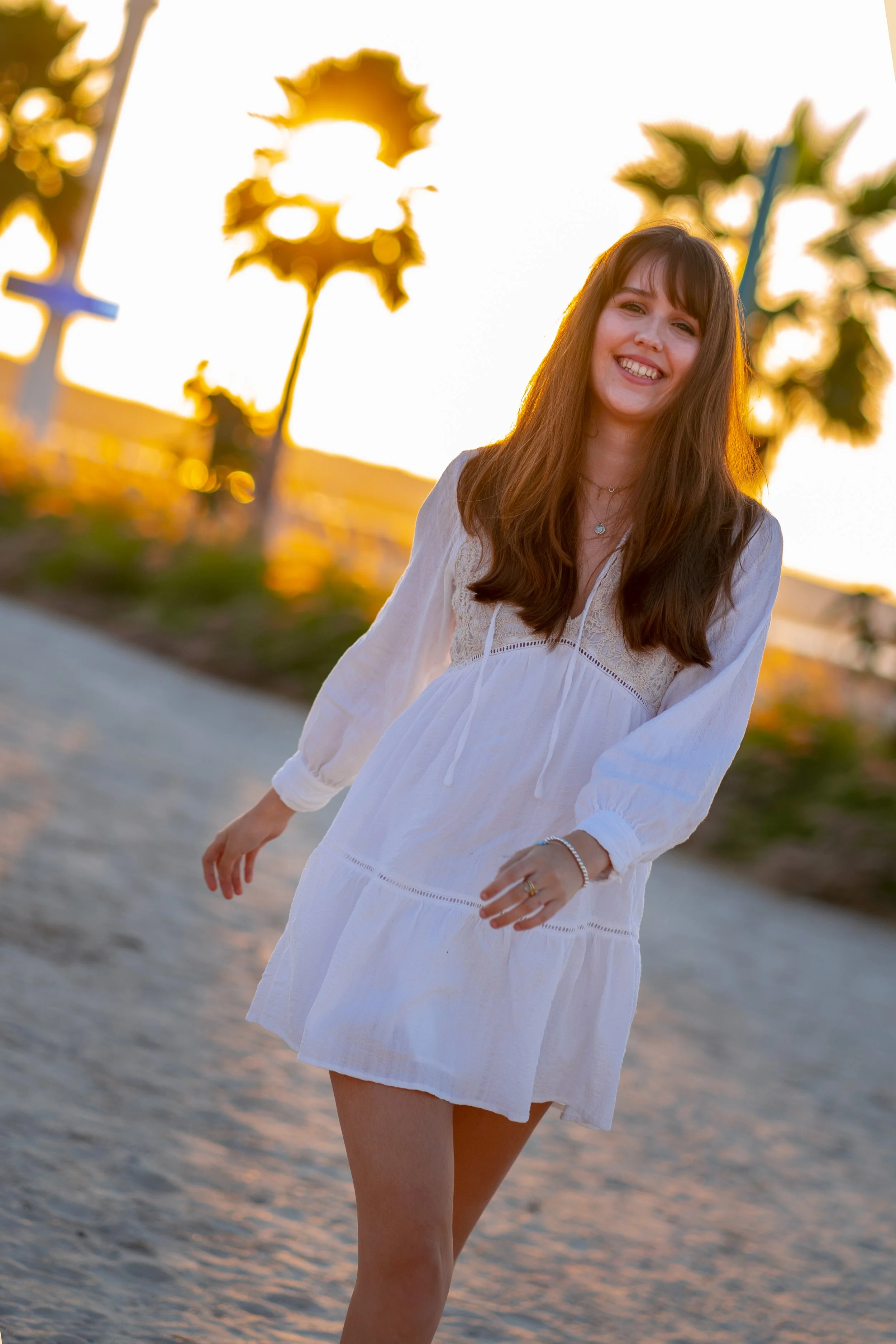 A young woman in a white dress walking along a beach at sunset, smiling.